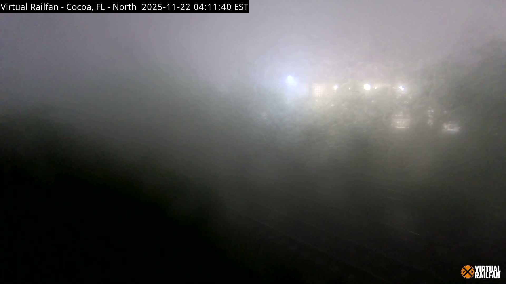 A very dense fog blankets an outdoor scene at night, almost completely obscuring distant buildings and streetlights, with faint dark lines in the foreground that could be railroad tracks.