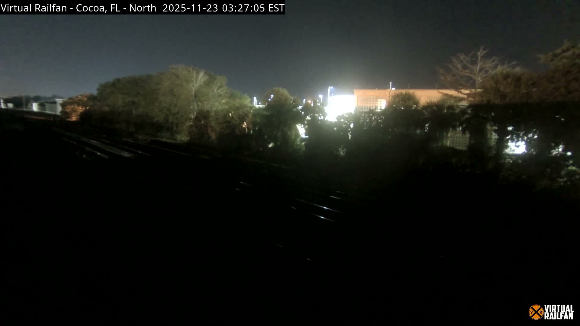 On a clear night, several dark railroad tracks occupy the foreground, leading towards a landscape of illuminated trees and buildings in the mid-ground, all under a dark sky.