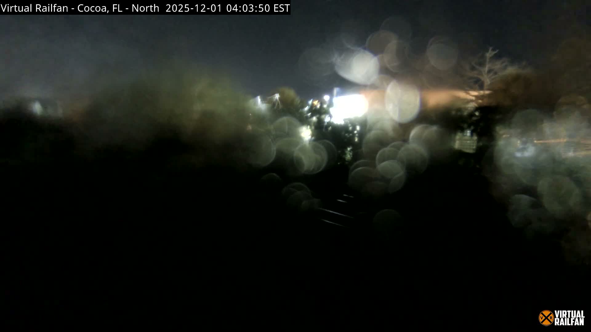 A dark, blurry outdoor scene at night is viewed through a rain-splattered lens, showing numerous bright water droplets and indistinct distant lights and shapes.