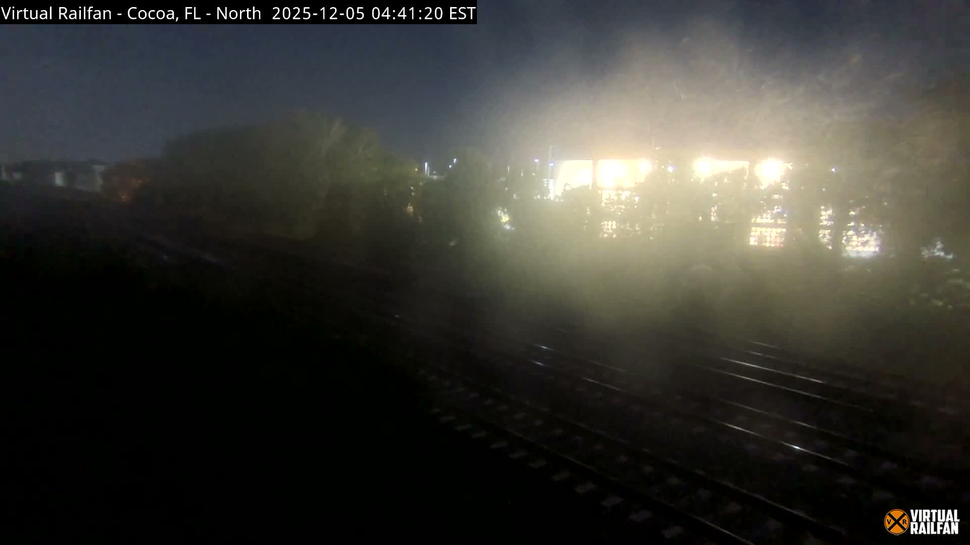 Dark train tracks curve into the foggy night, with diffused bright lights illuminating the dense mist in the background.