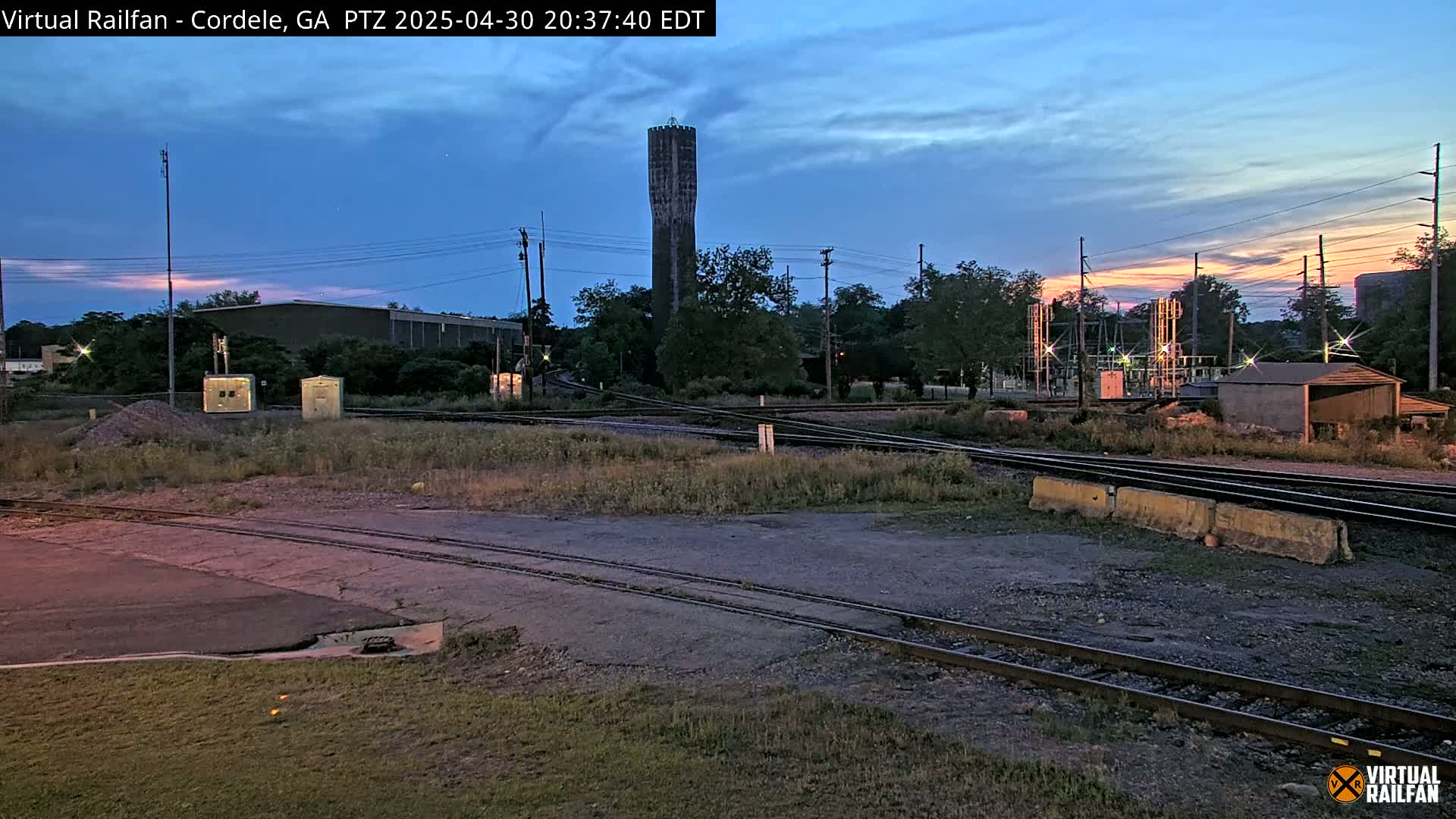 A tall, slender water tower stands in the distance under a twilight sky, near a railway intersection and industrial buildings.