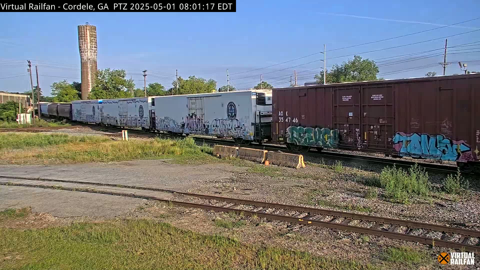 A freight train with graffiti-covered cars passes by under a clear sky near a tall water tower.