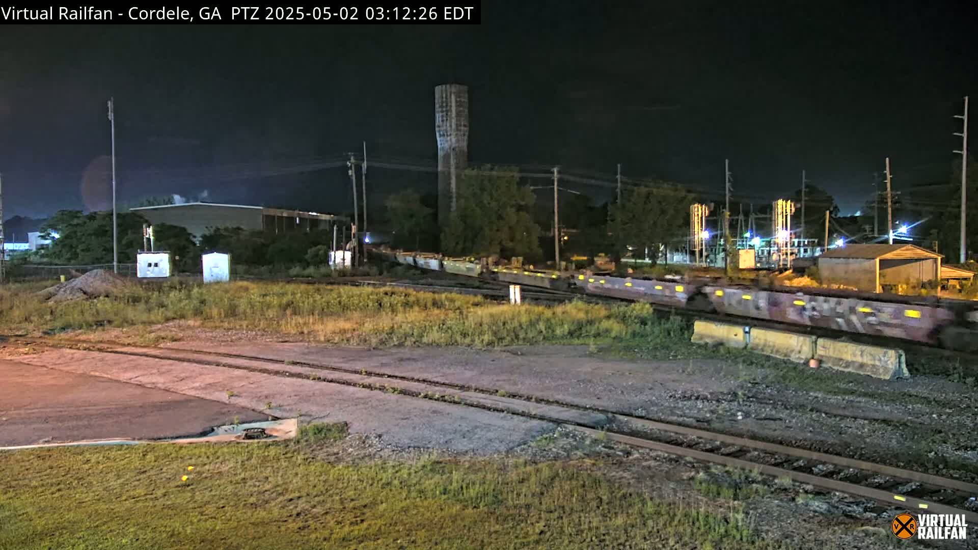 At night, a long freight train moves past a water tower and industrial buildings under a dark sky.
