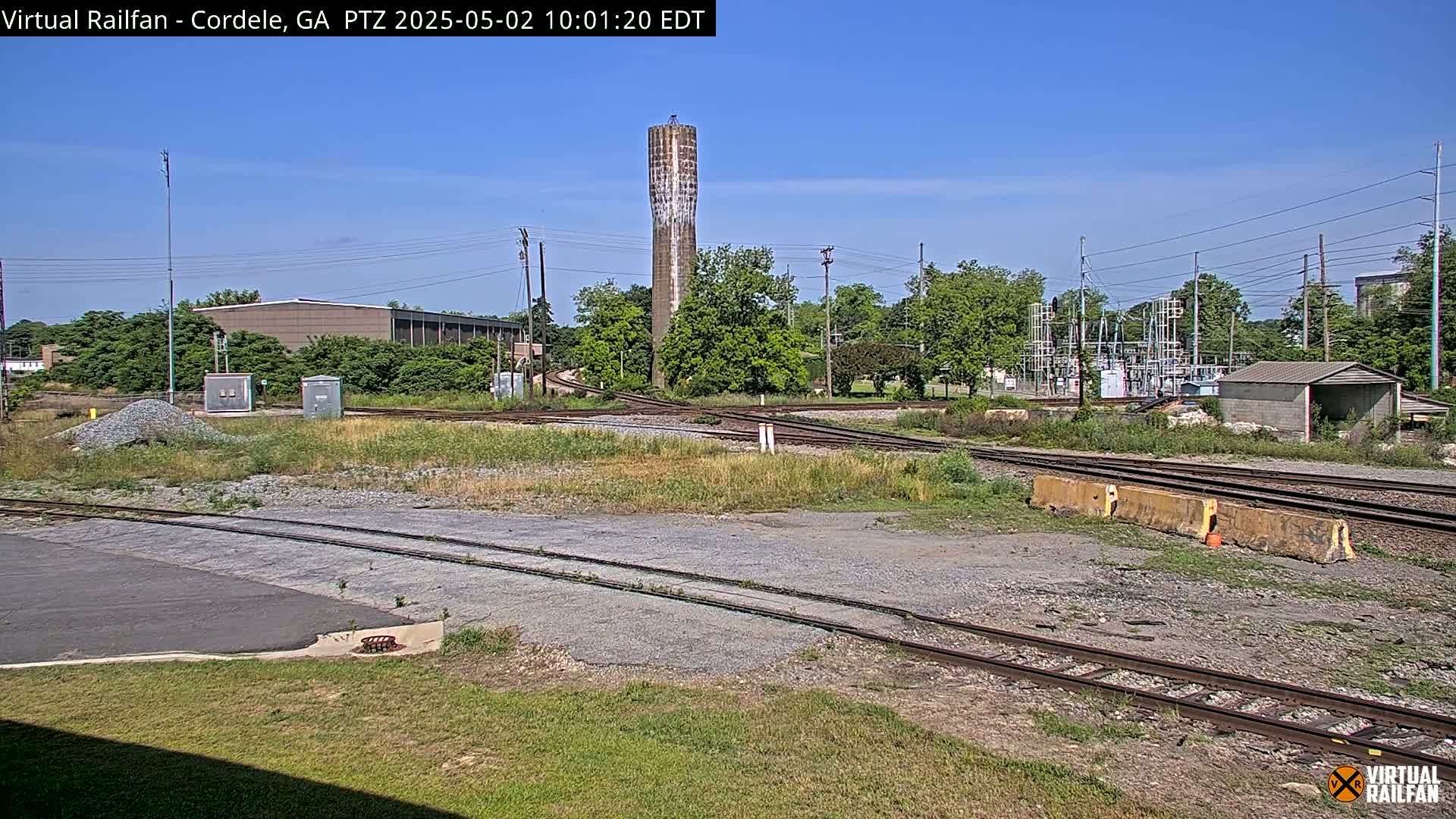 A railroad yard with converging tracks under a clear blue sky, featuring a tall water tower and surrounding vegetation.