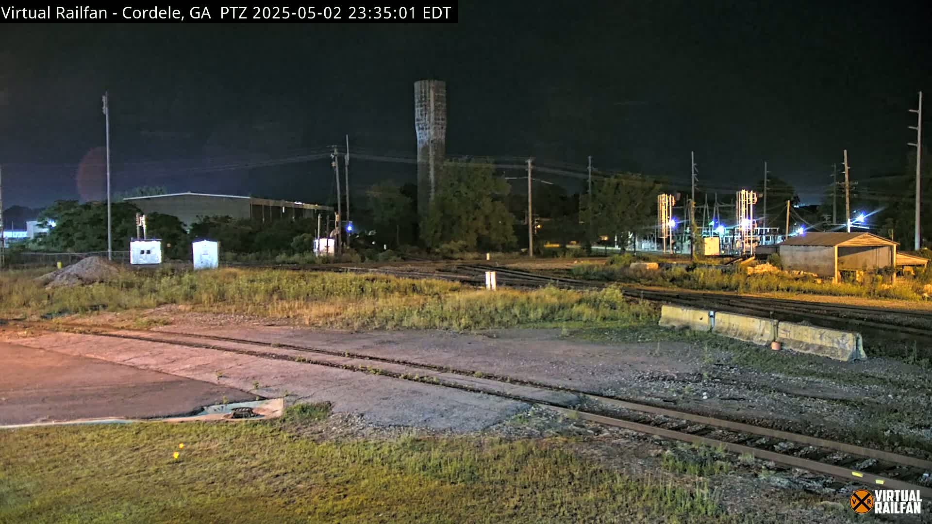 A nighttime view of a rail yard with a tall tower, buildings, and overgrown vegetation under a dark sky.