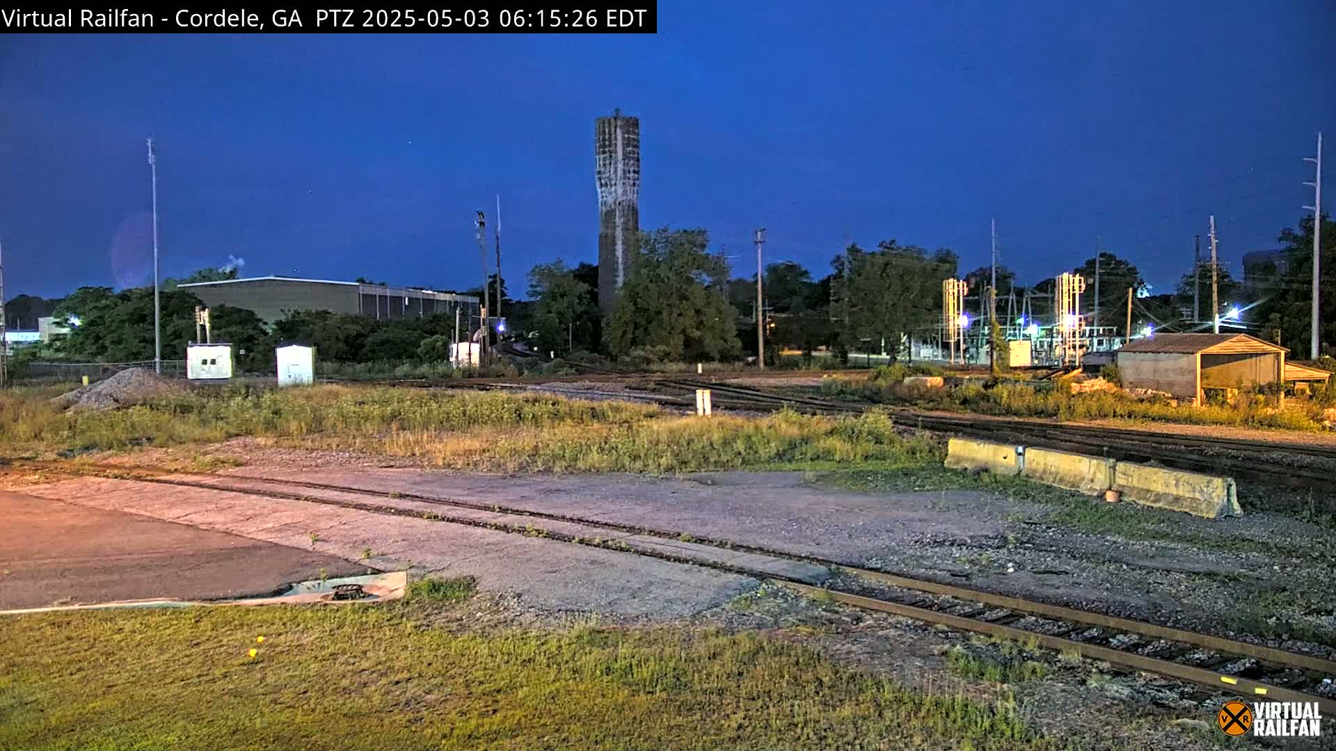 Under a twilight sky, a train yard with a tall, narrow tower is visible, alongside various industrial buildings and railway tracks.