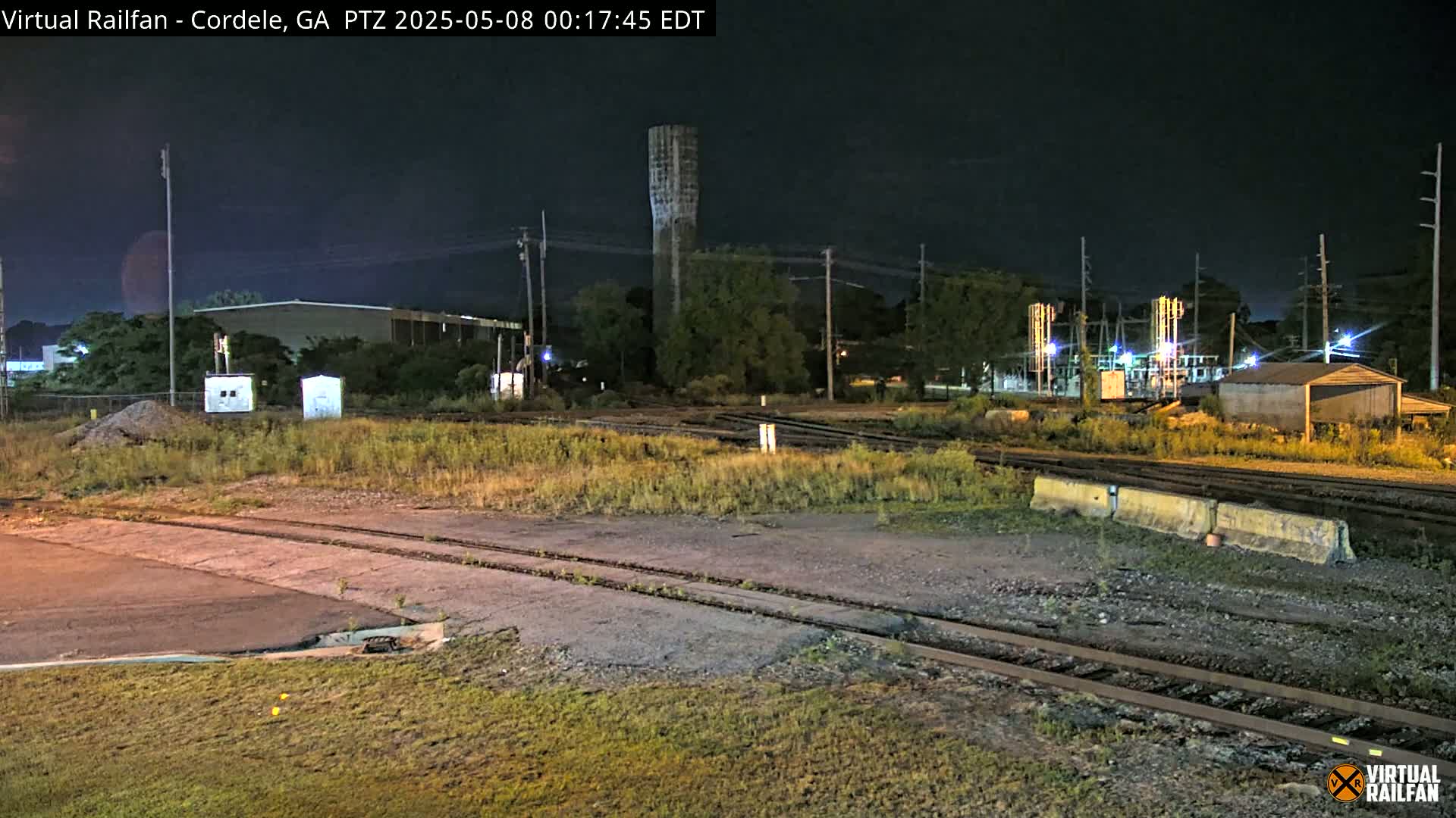 A nighttime view of railroad tracks and industrial buildings under a dark sky, with a tall, thin tower visible in the background.