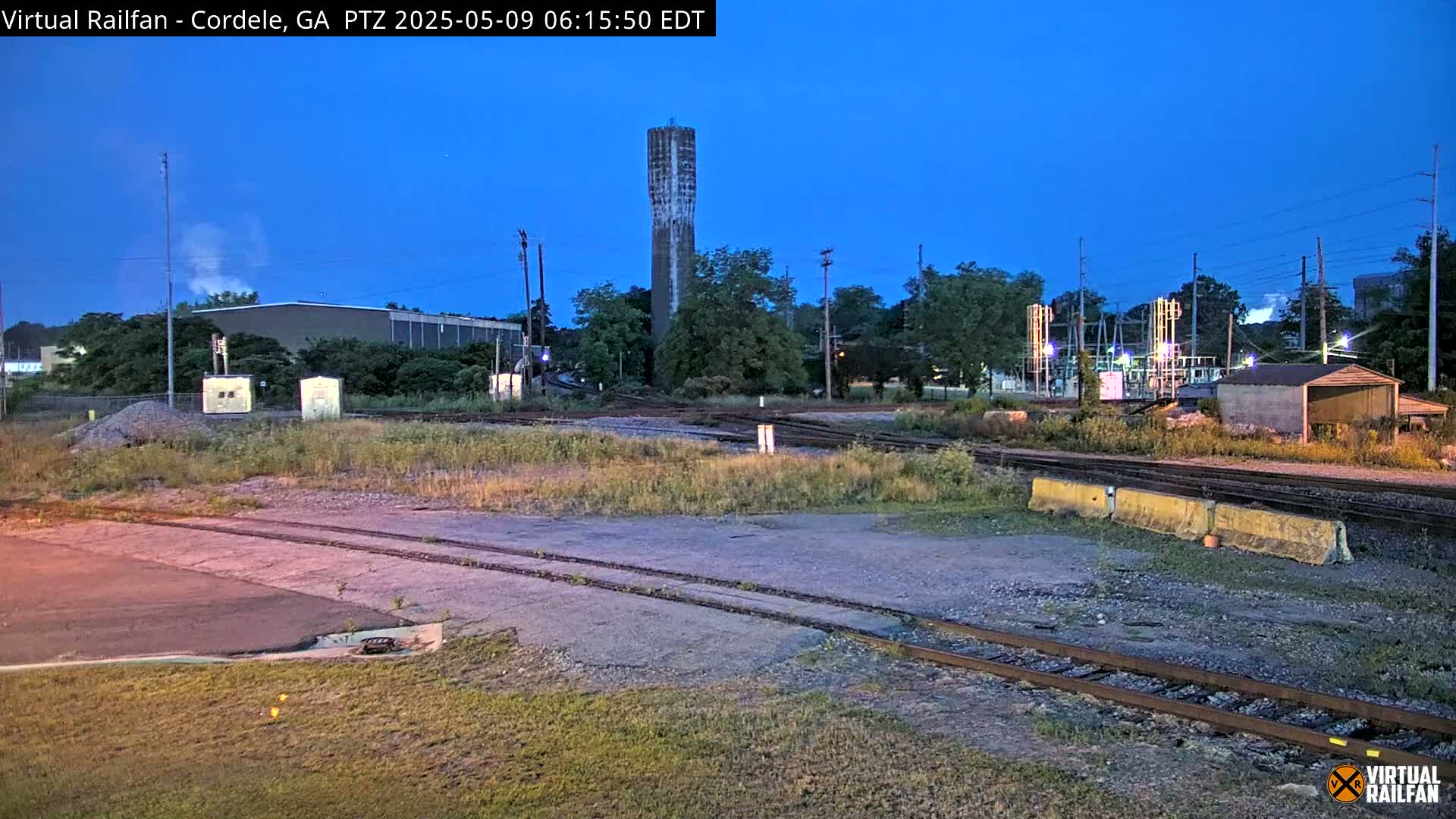 Under a twilight sky, a railway yard with a tall water tower is surrounded by low vegetation, buildings, and electrical equipment.