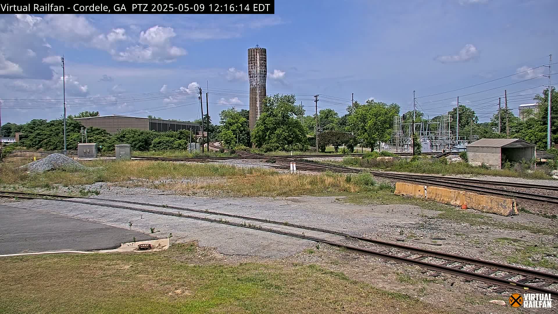 A train yard with a tall, slender water tower in the background under a partly cloudy sky.