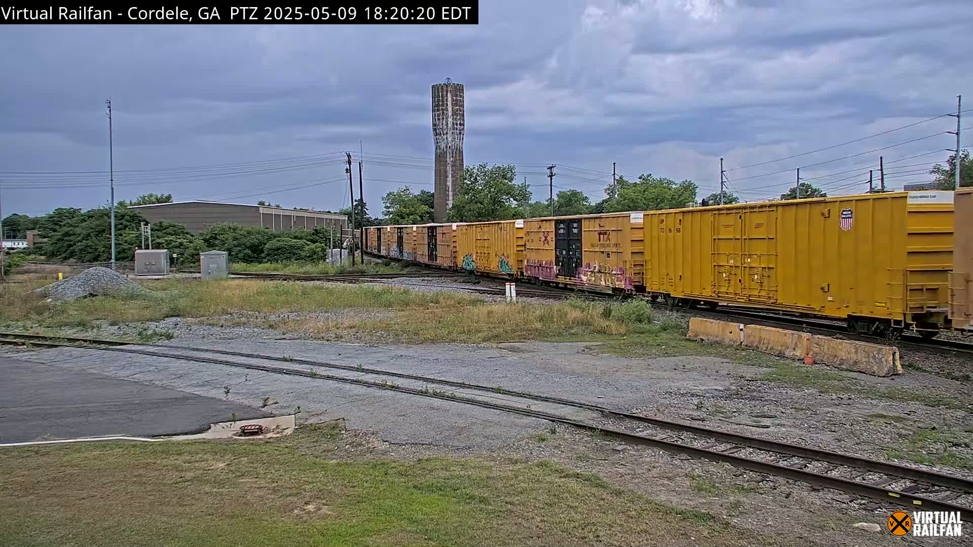 A long freight train of yellow boxcars passes by under a cloudy sky near a water tower and industrial buildings.