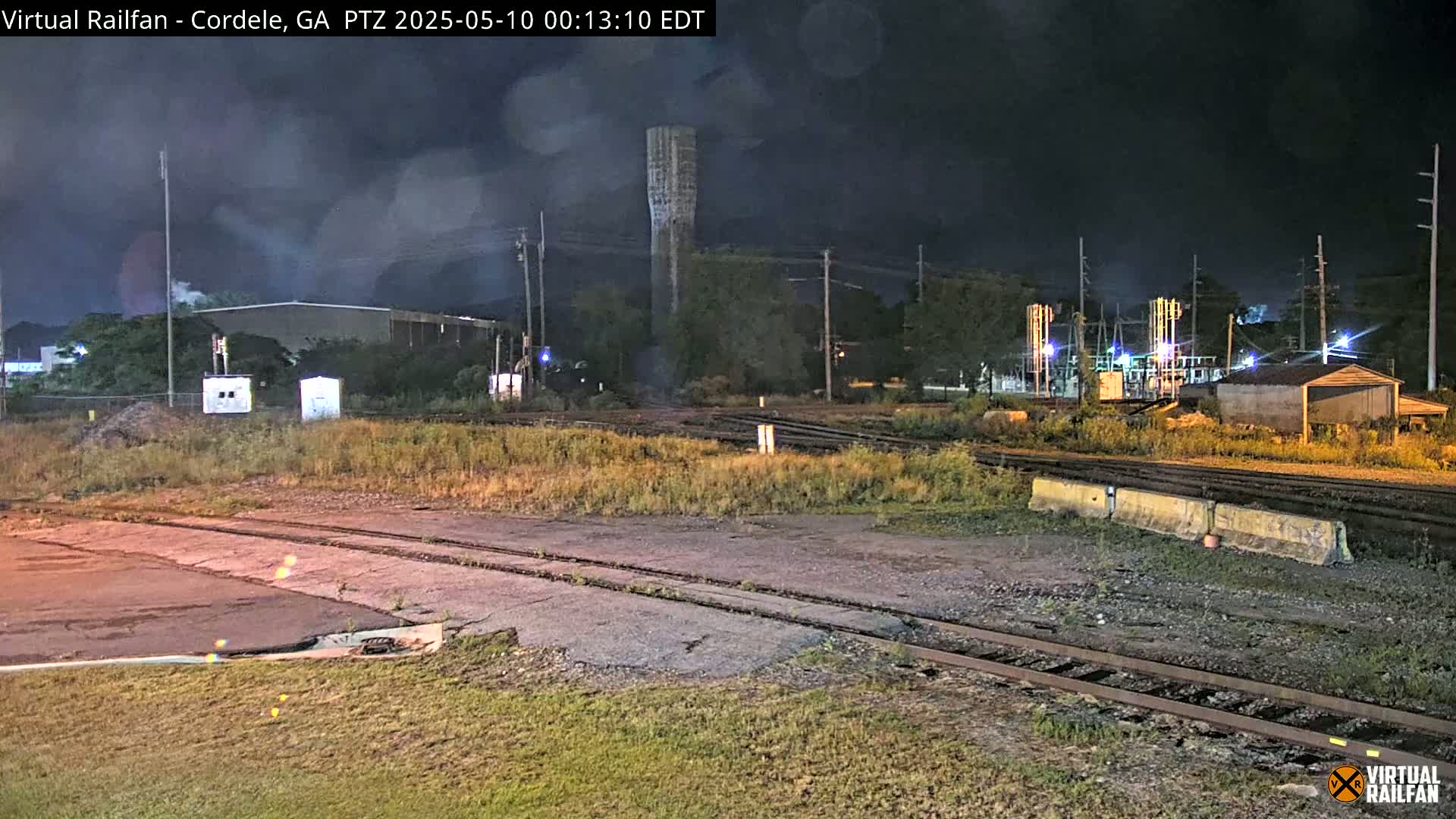A nighttime view of a rail yard with a tall water tower in the background under a dark, cloudy sky.