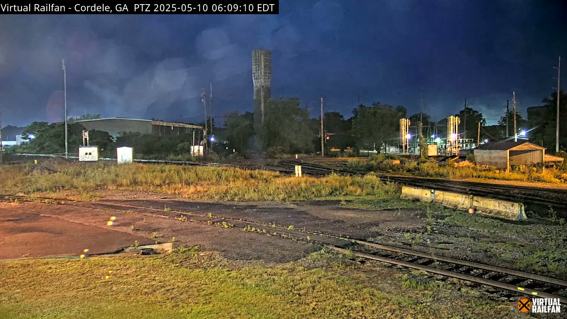 Under a dark, mostly clear night sky, a nighttime view shows railroad tracks and industrial buildings, including a tall, slender structure that appears to be a water tower.