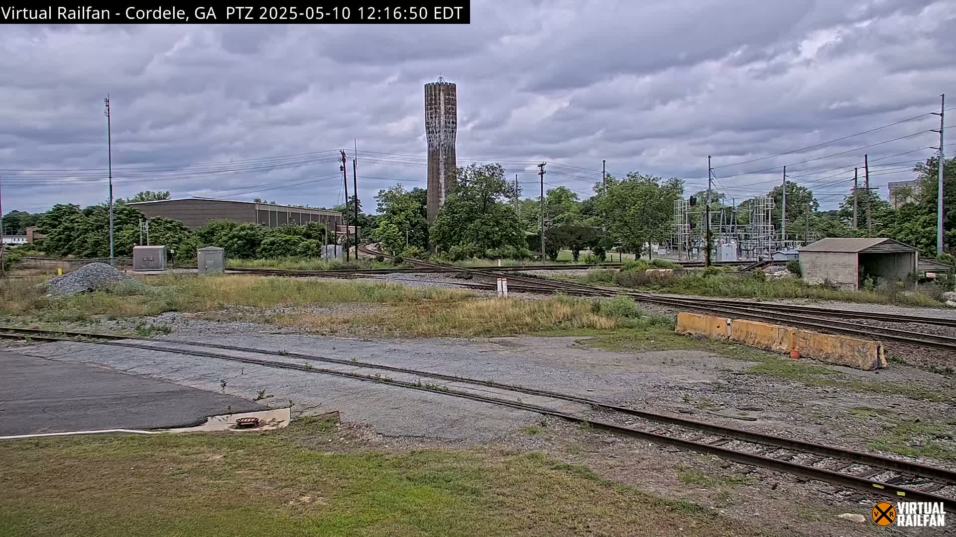 Under a cloudy sky, a view of railroad tracks and switches with a water tower in the background, surrounded by industrial buildings and vegetation.