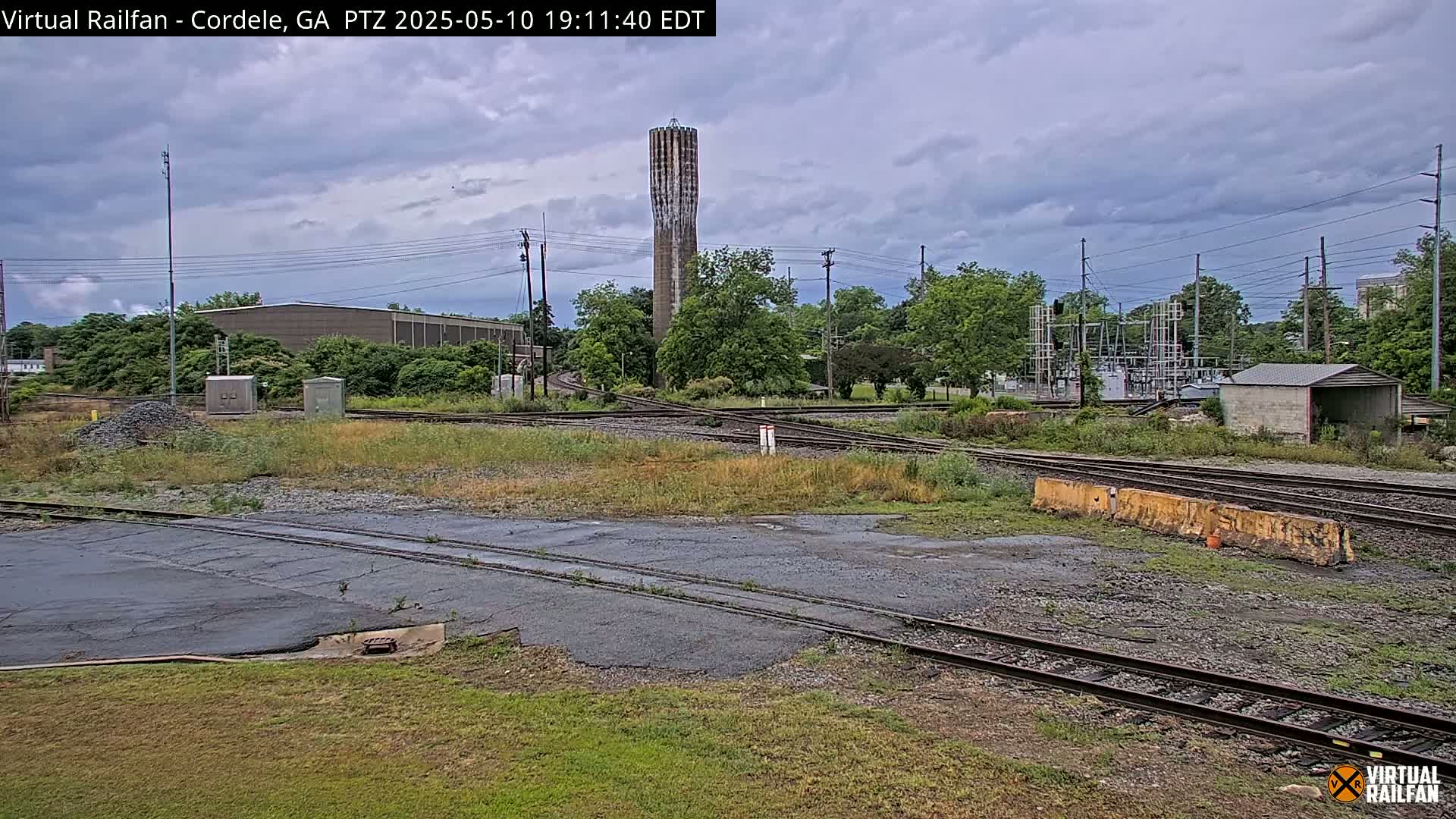 A tall, slender water tower is visible in the distance beyond a railway switching yard under a mostly cloudy sky.