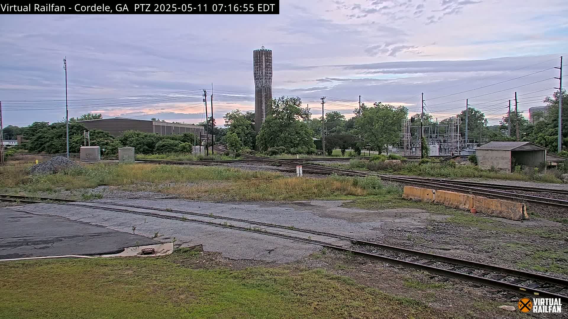 A tall water tower stands in the distance behind a railroad yard with multiple tracks converging under a mostly cloudy sky.