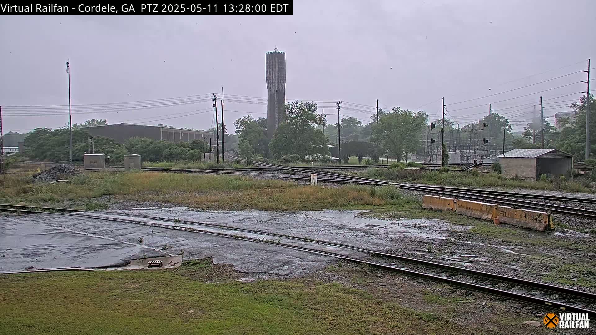 A rain-soaked railroad crossing with a tall water tower in the background, surrounded by industrial buildings and greenery.