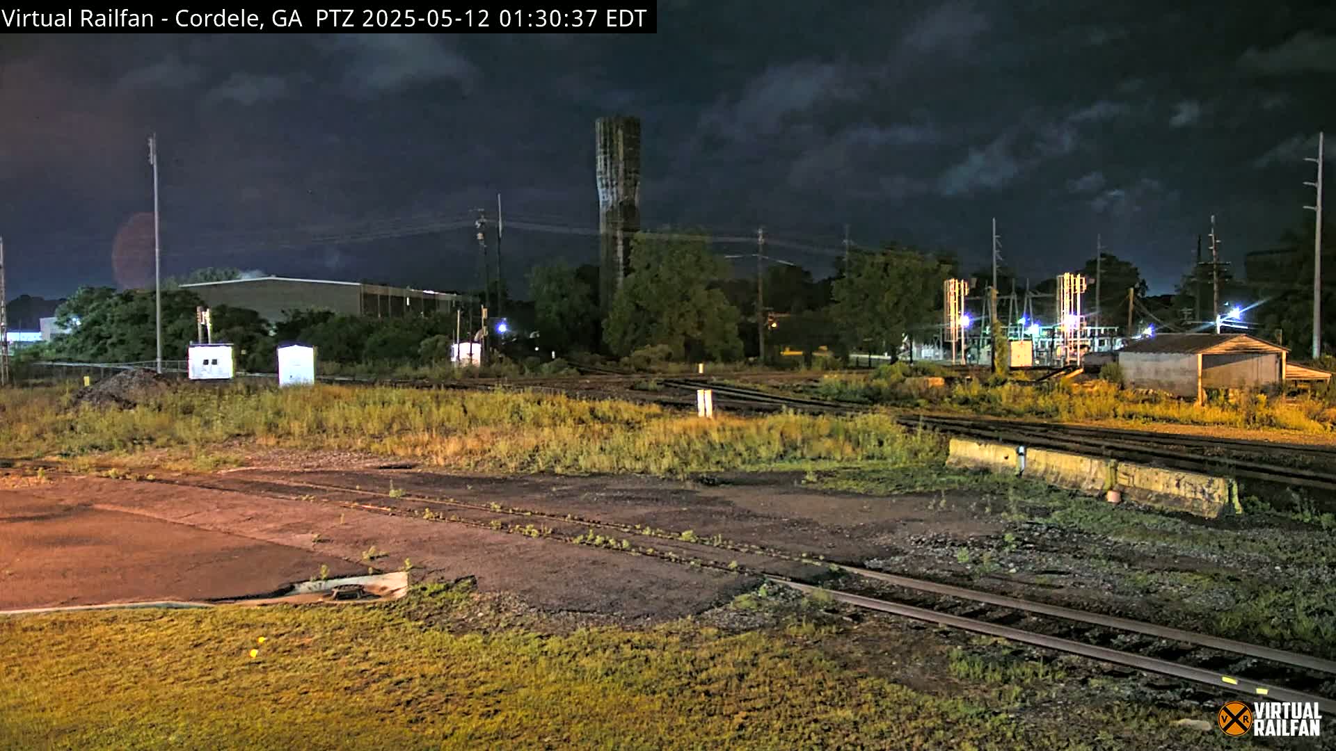 Under a partly cloudy night sky, railroad tracks and switching yards are visible near some industrial buildings and overgrown vegetation.