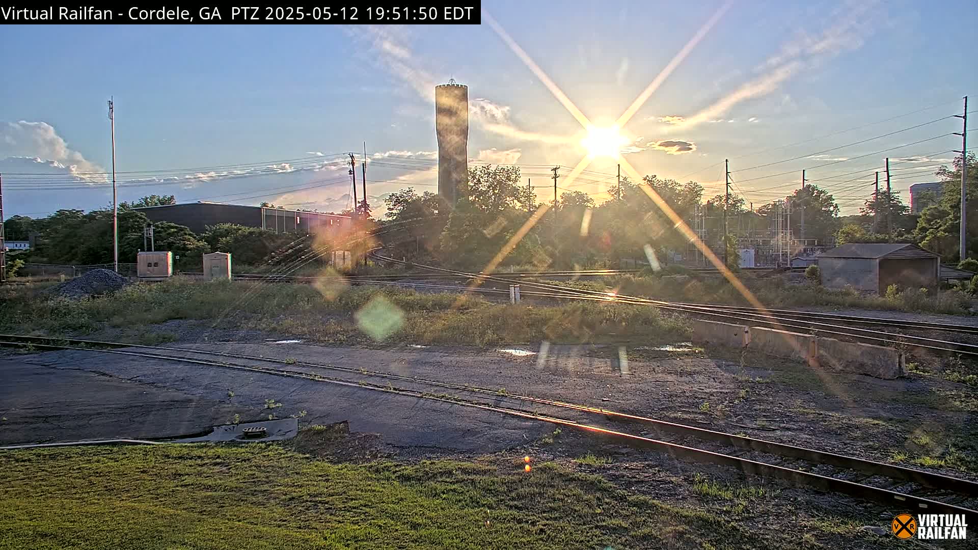 A sunset illuminates a railroad yard with intersecting tracks, a water tower in the distance, and some low-lying buildings and vegetation.