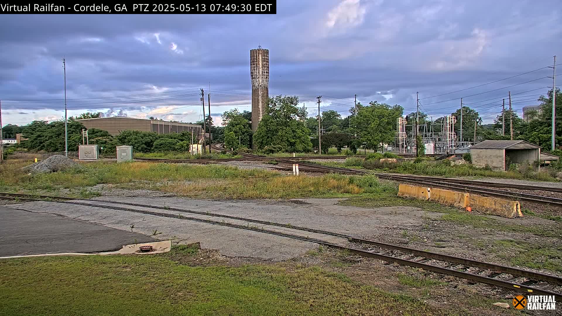 A railway switching yard with a tall, slender water tower in the background under a partly cloudy sky.