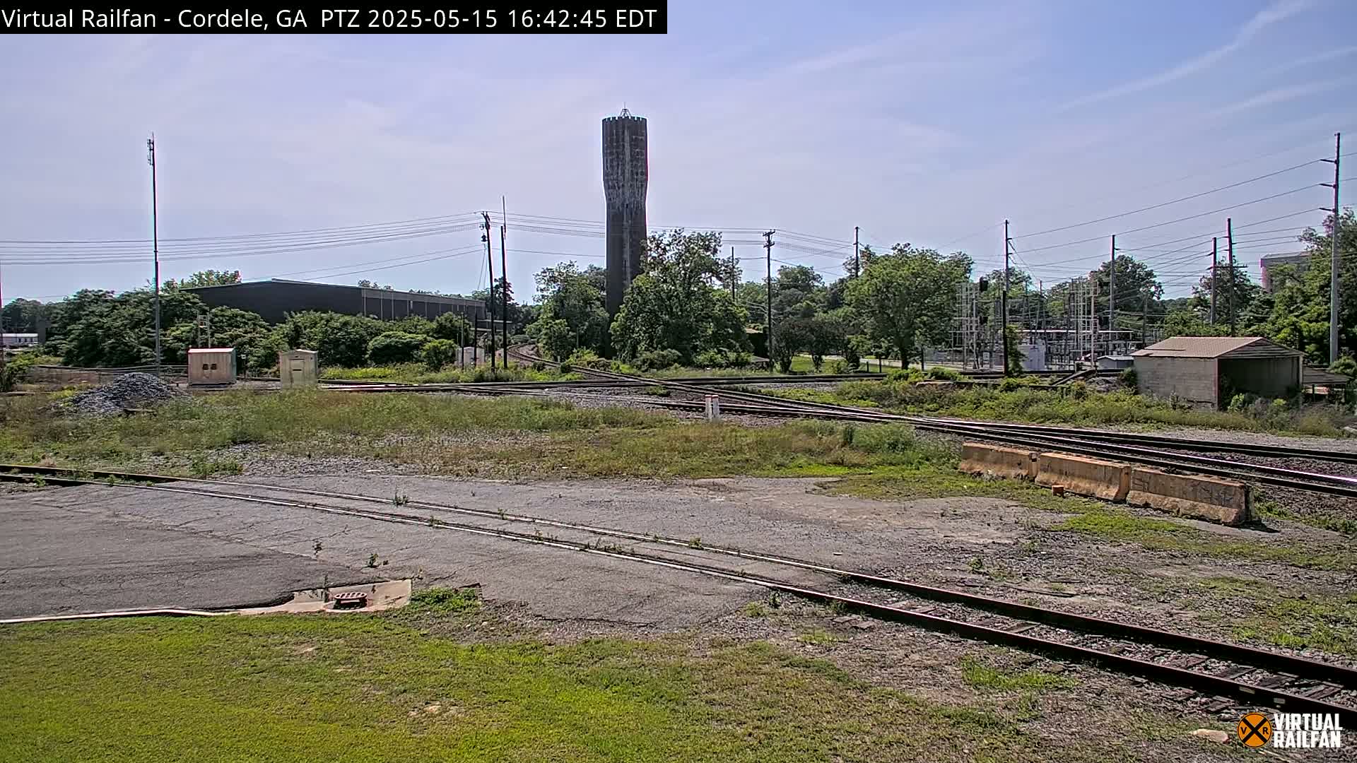 Under a mostly clear sky, a tall water tower stands in the background of a railway switching yard with multiple tracks and some small buildings.