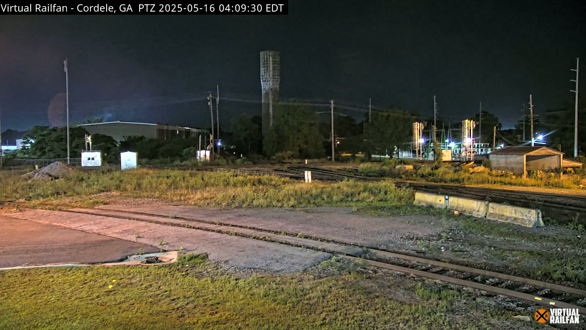 At night, a nighttime view shows railroad tracks, a tall water tower, some industrial buildings, and overgrown vegetation.