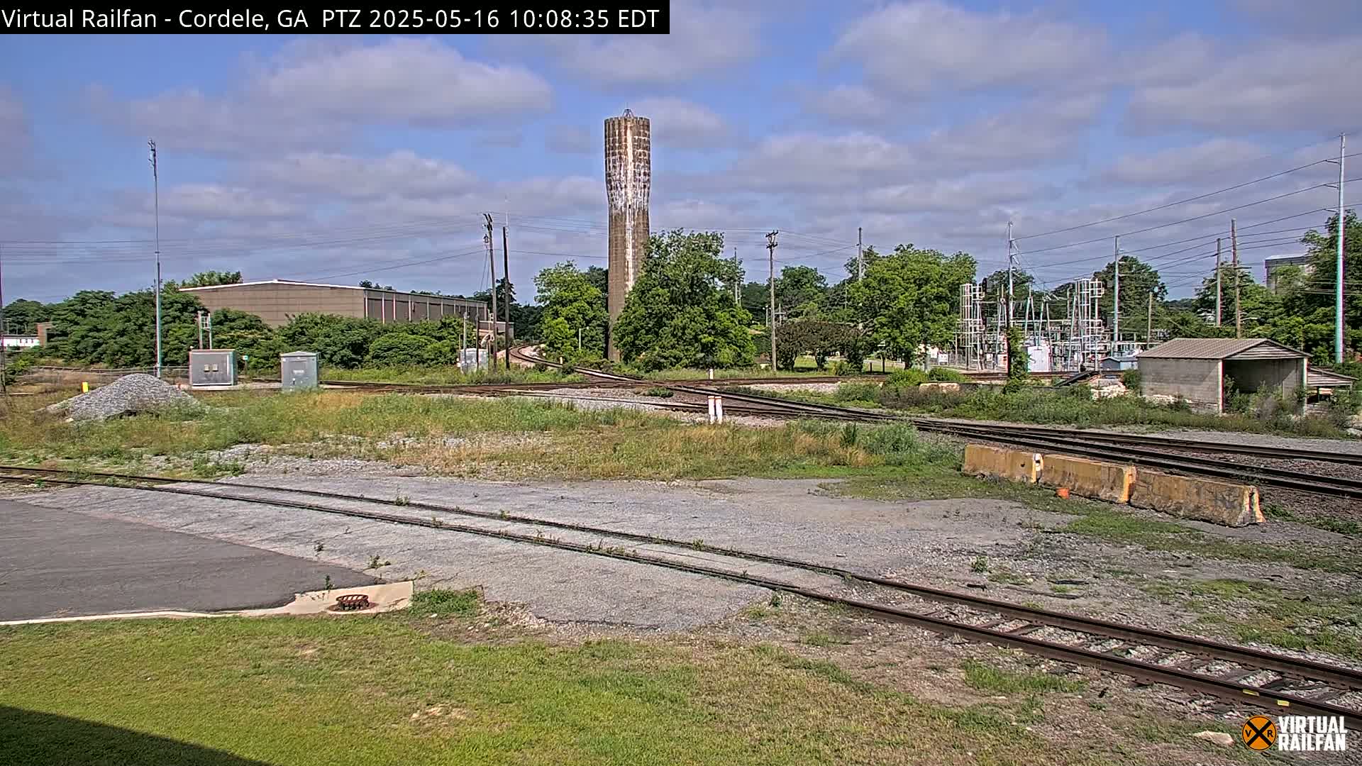 A partly cloudy day shows a rail yard with multiple tracks converging near a tall, slender water tower and industrial buildings, surrounded by green vegetation.
