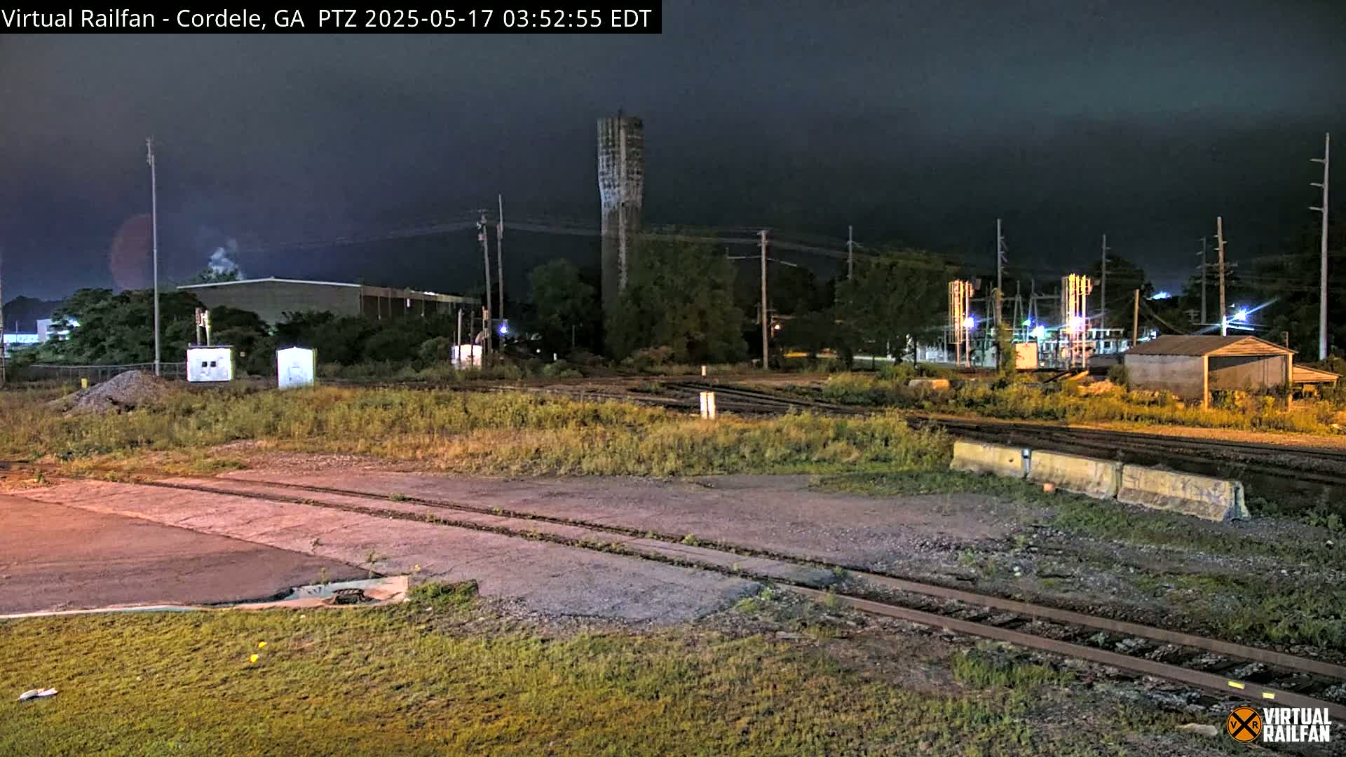 A nighttime view of a rail yard with a tall tower, industrial buildings, and overgrown vegetation under a dark sky.