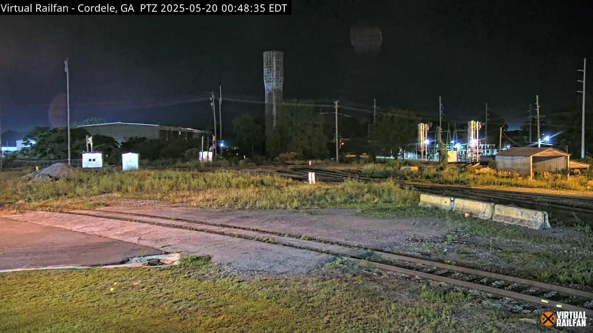 A nighttime view of railroad tracks and industrial buildings under a dark sky, with a tall water tower visible in the background.