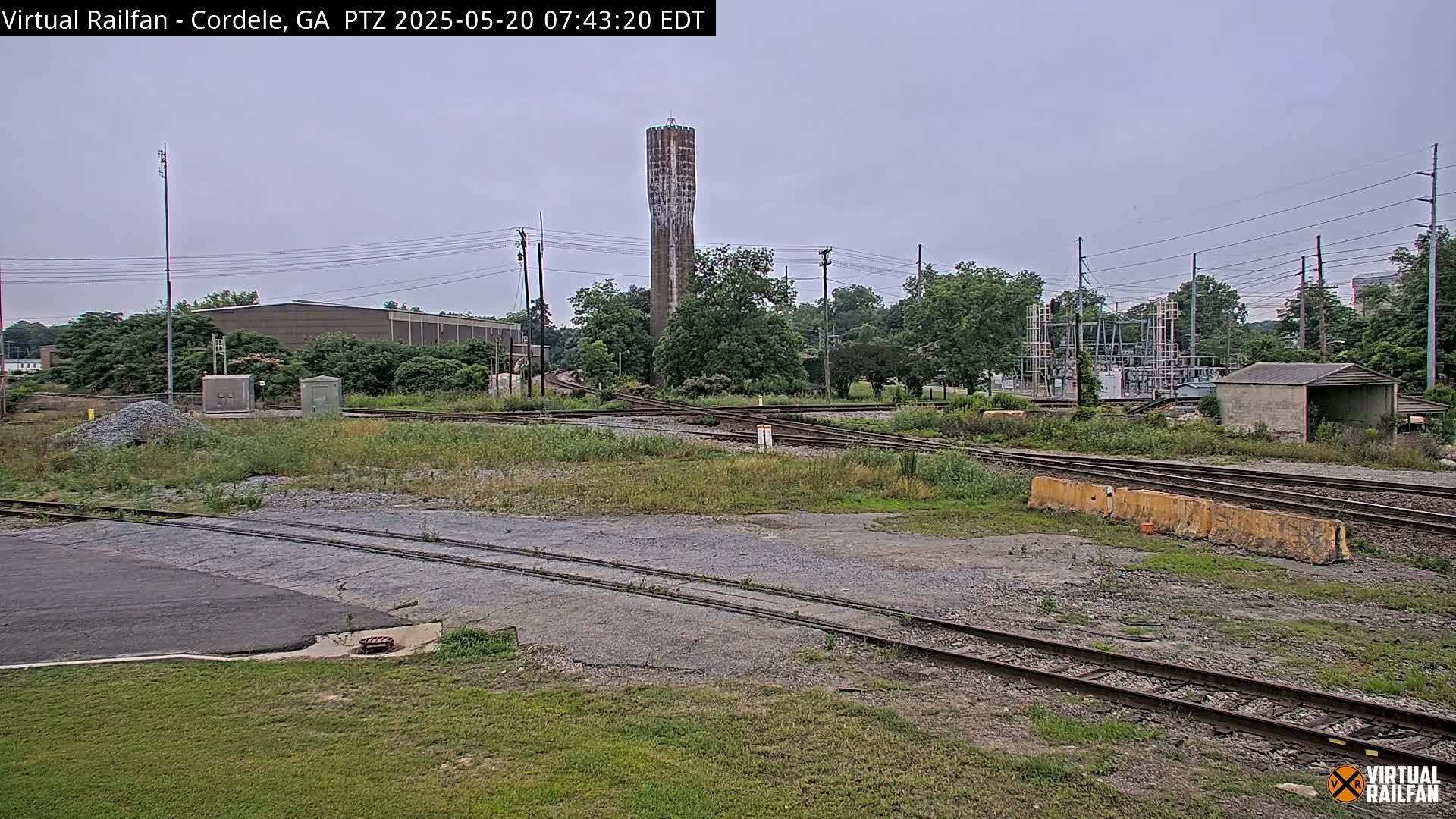 A water tower stands in the background of an overcast day, overlooking a railway switching yard with various buildings and overgrown vegetation.