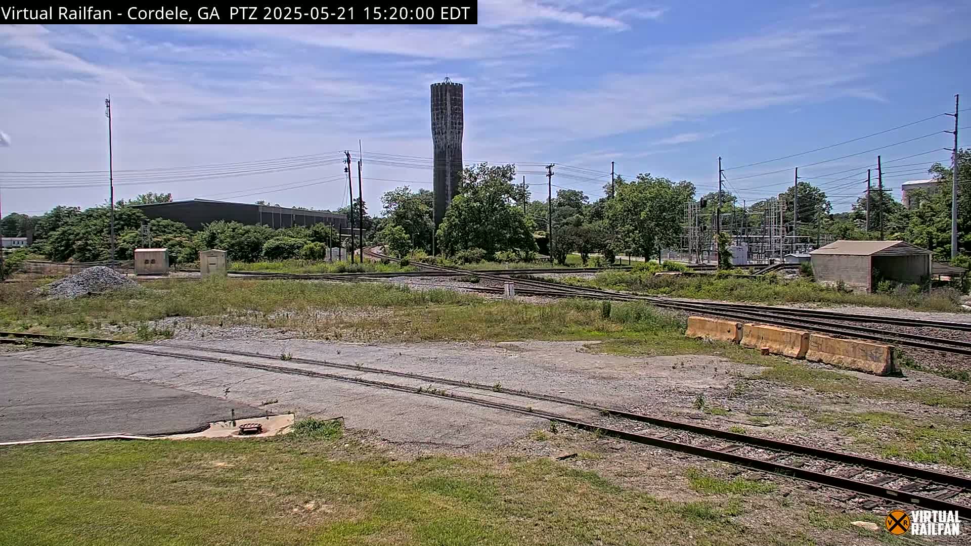A water tower stands in the background of a sunny outdoor scene featuring a railway switching yard with overgrown vegetation.