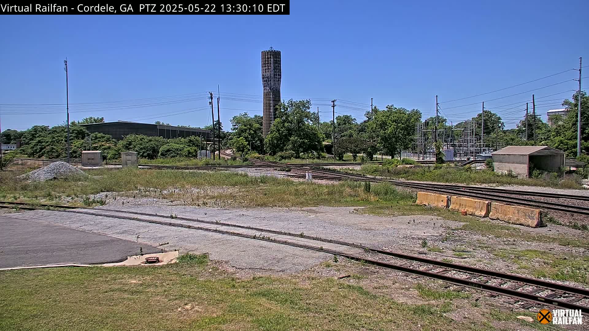 A tall, slender water tower stands in the background of a sunny scene featuring a railway switching yard with several tracks, surrounded by trees and industrial buildings.