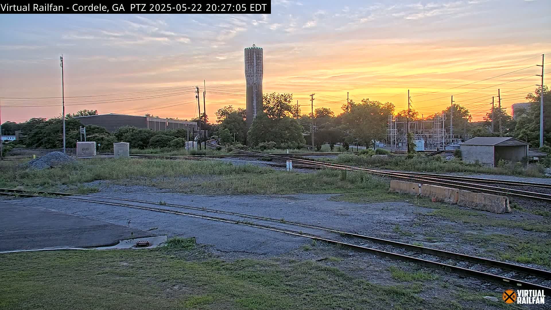 A tall, slender water tower stands in the background of a sunset scene, overlooking a railway yard with multiple tracks, some overgrown weeds, and a small concrete building.