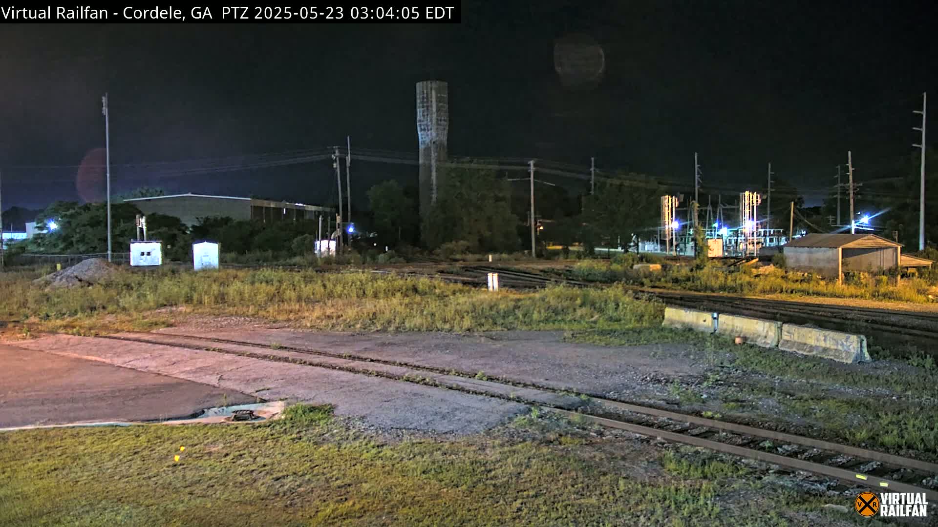 At night, a train yard with a water tower and various industrial buildings is shown under a clear, dark sky.
