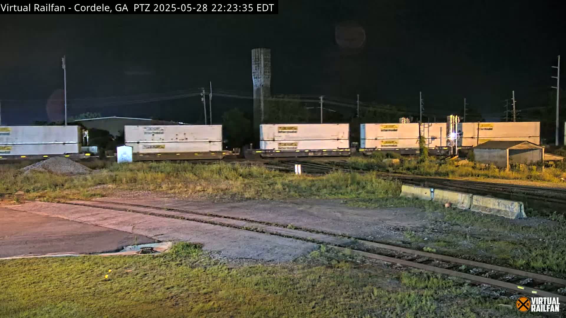 At night, a train carrying several white shipping containers sits stationary on railroad tracks beside a weedy area.