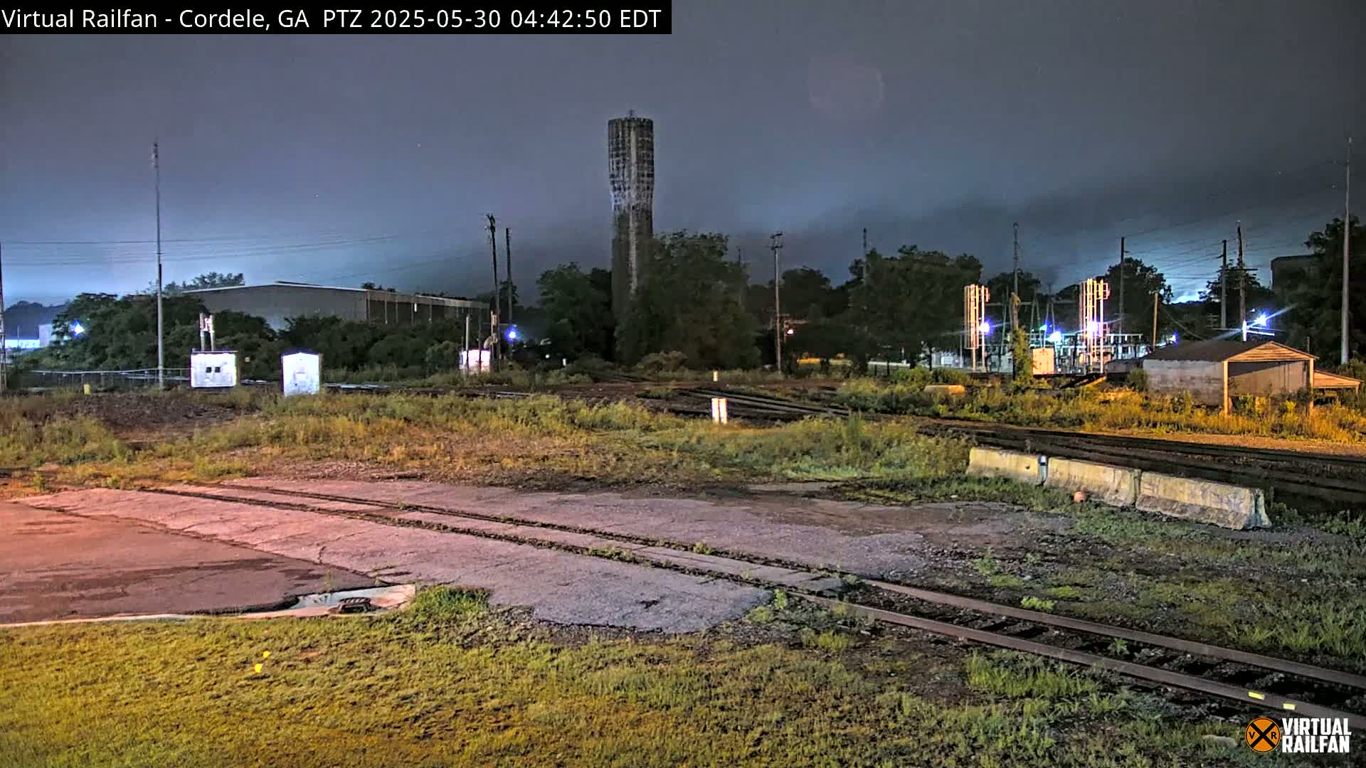 A nighttime view of a railway yard with a tall water tower in the background under a cloudy sky.