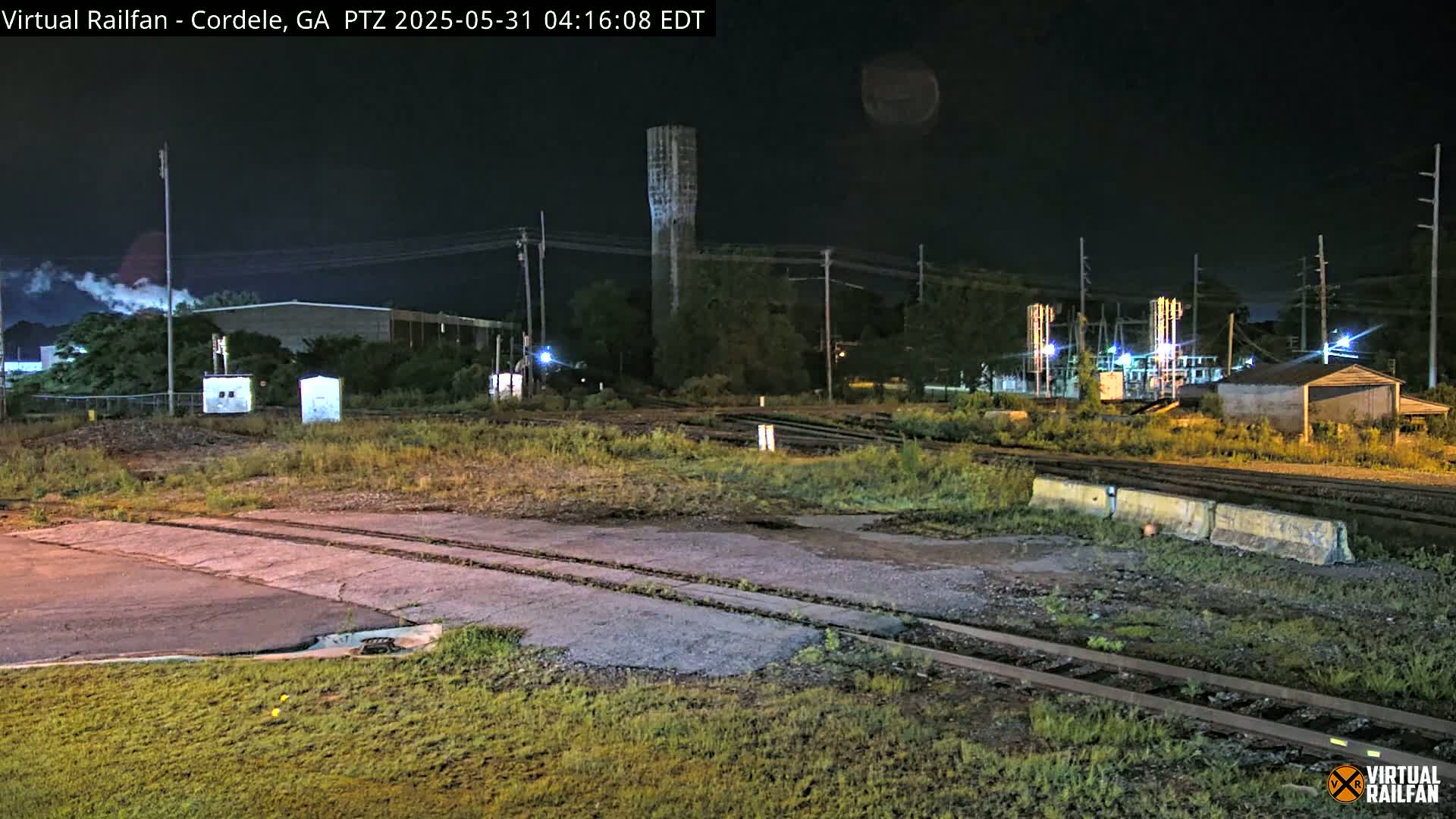 A nighttime view of a rail yard with a tall tower, industrial buildings, and overgrown vegetation under a clear, dark sky.