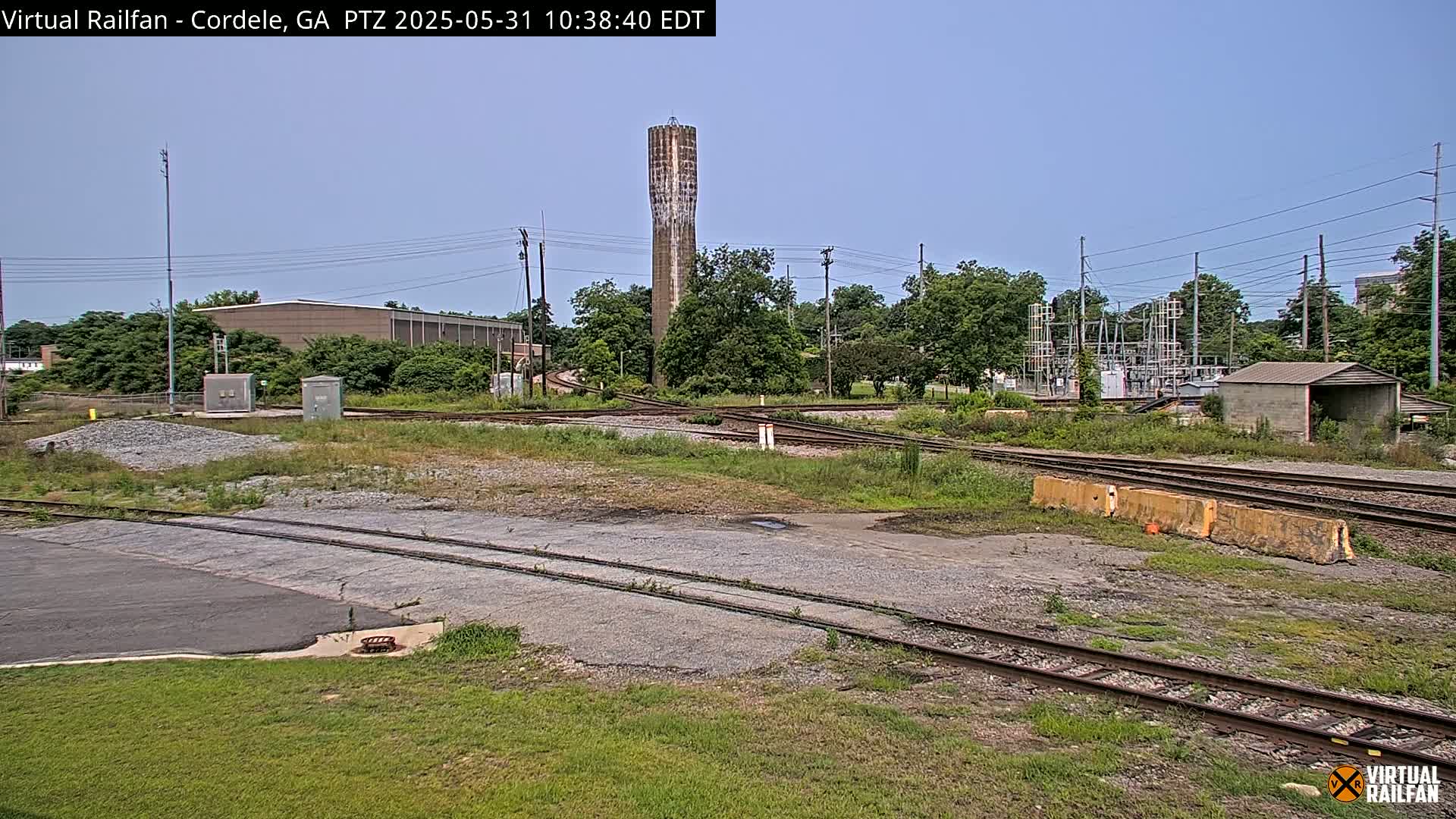 A tall, slender water tower stands in the background of a sunny scene featuring a railway switching yard with several tracks, surrounded by greenery and industrial buildings.