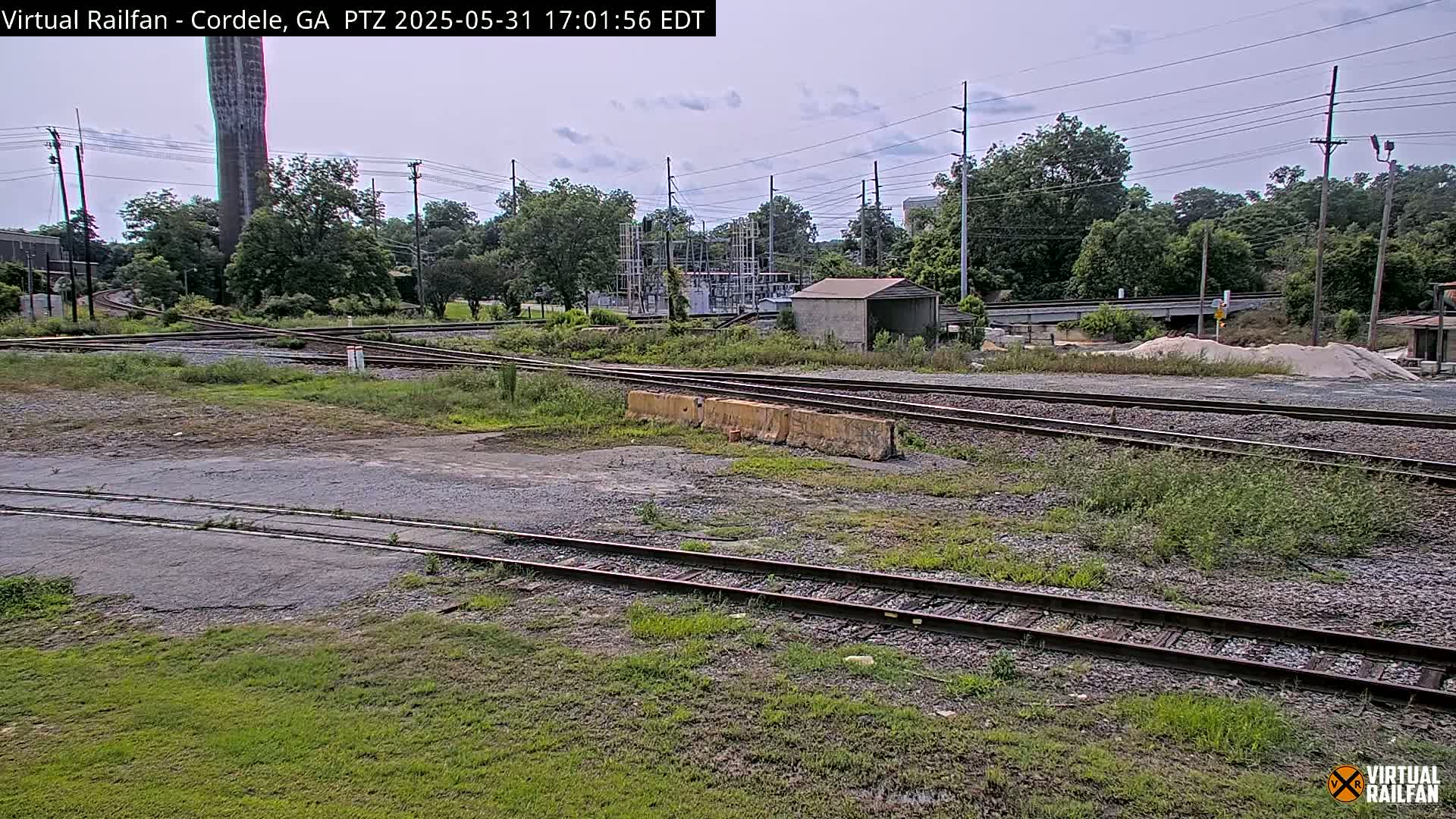 A railway switching yard with several sets of tracks, overgrown weeds, a concrete structure, and trees under a partly cloudy sky.