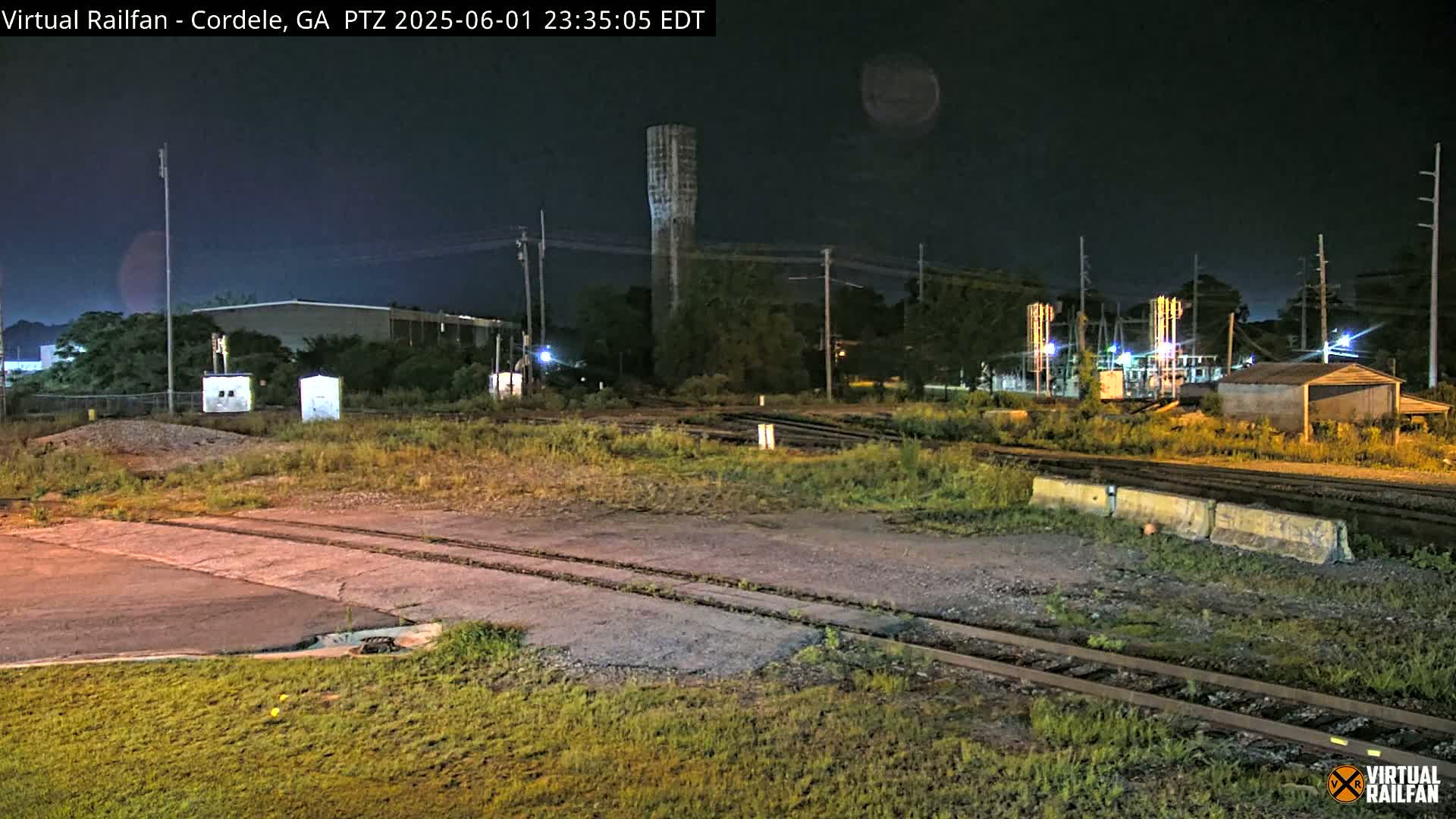 A nighttime view of a rail yard with a tall tower, several buildings, and overgrown vegetation under a dark, clear sky.