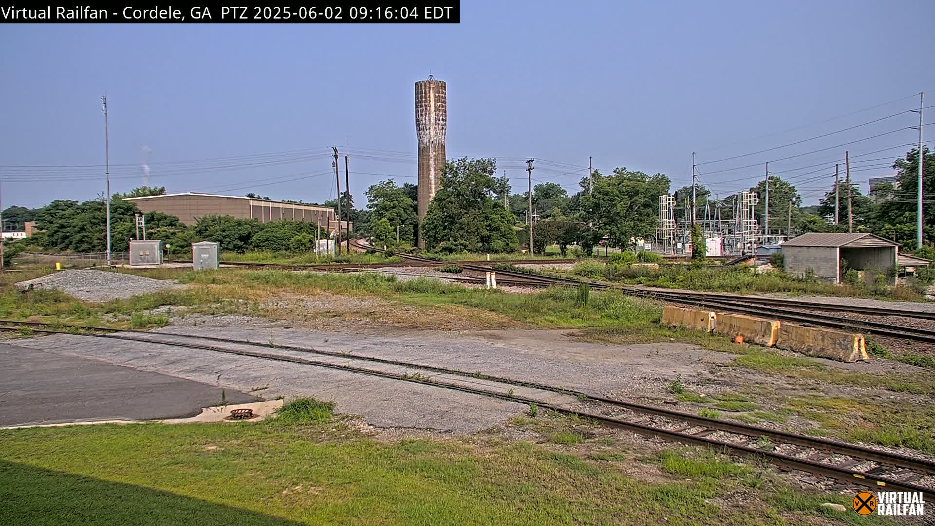 A view of a railroad switching yard on a sunny day, featuring a tall water tower in the background and various industrial buildings and structures.