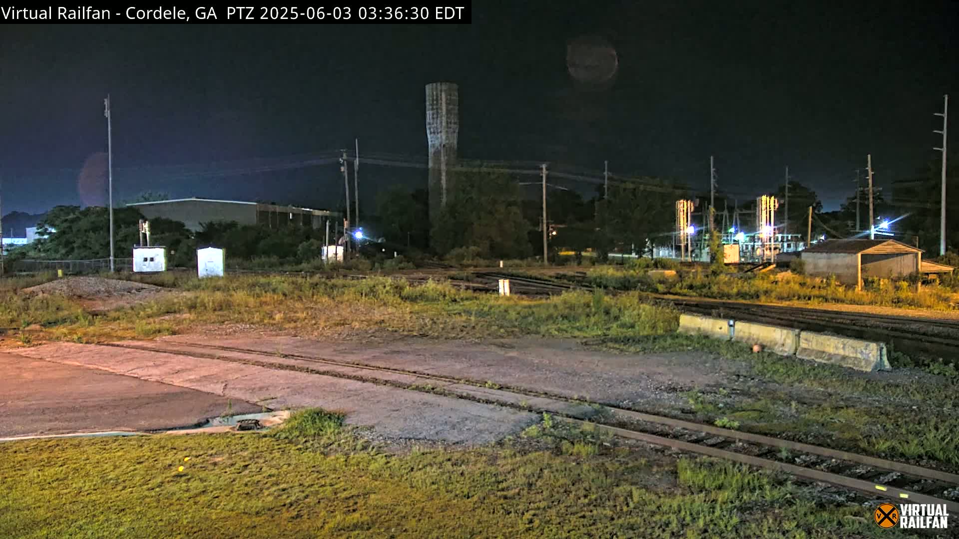 At night, a water tower and industrial buildings are visible near railroad tracks and overgrown vegetation under a dark sky.