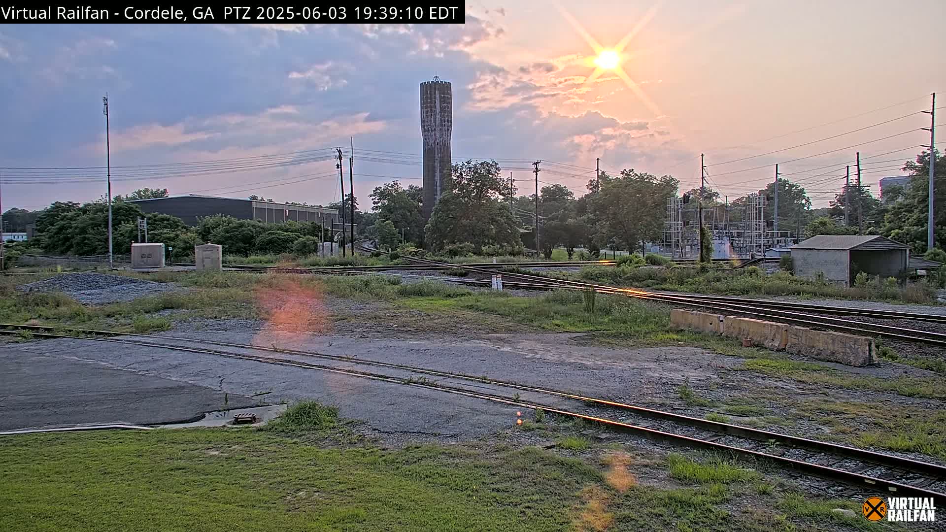 A sunset illuminates a rail yard with intersecting tracks, a tall water tower, and surrounding industrial buildings and vegetation.