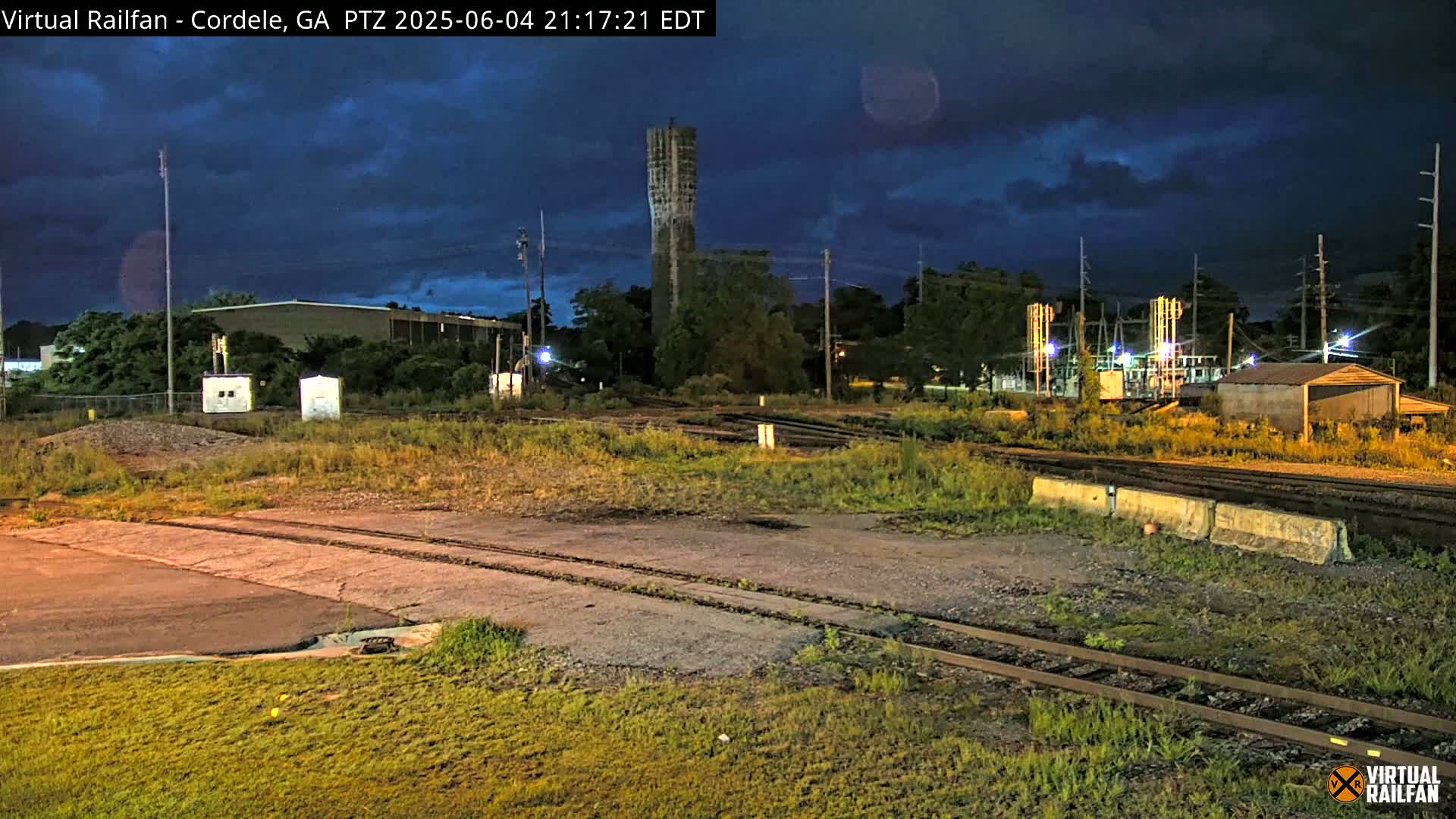 Under a partly cloudy night sky, a nighttime view shows railroad tracks, industrial buildings, and a tall, slender tower.