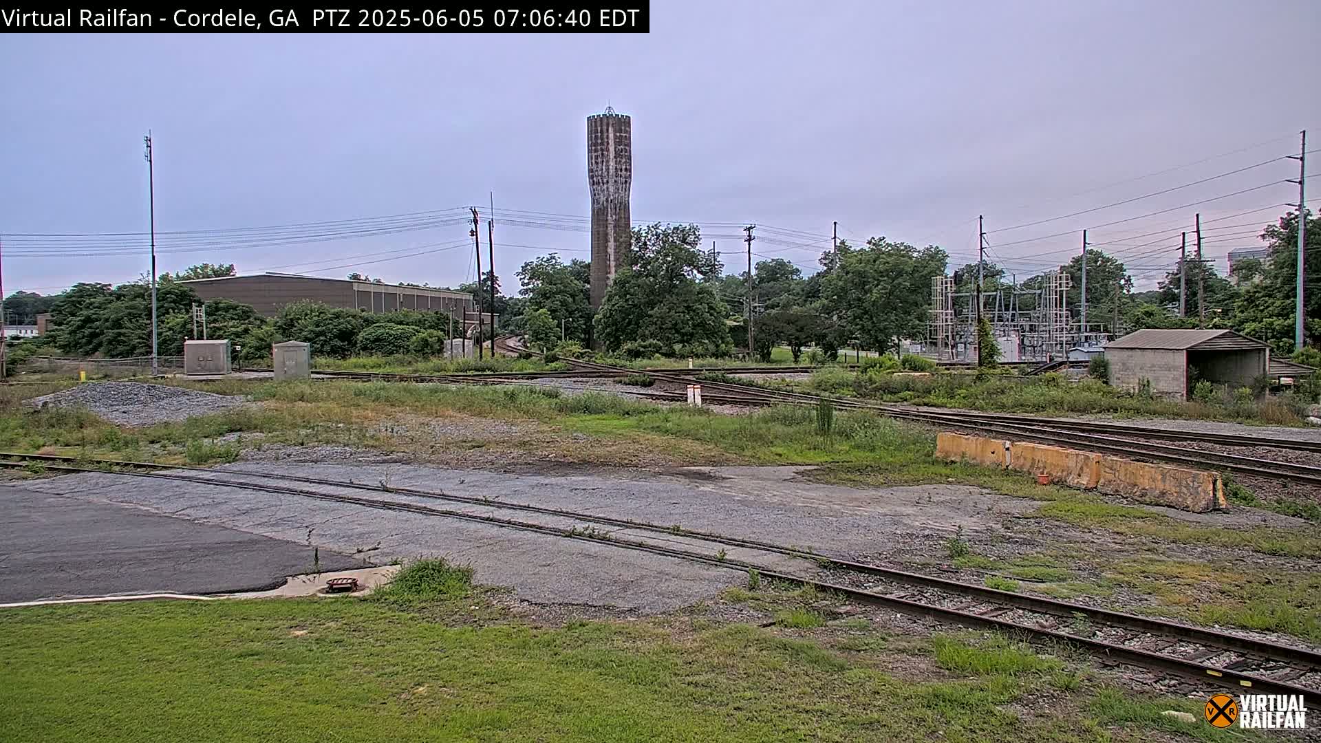 A tall, slender water tower stands in the distance behind a railway switching yard on an overcast day.