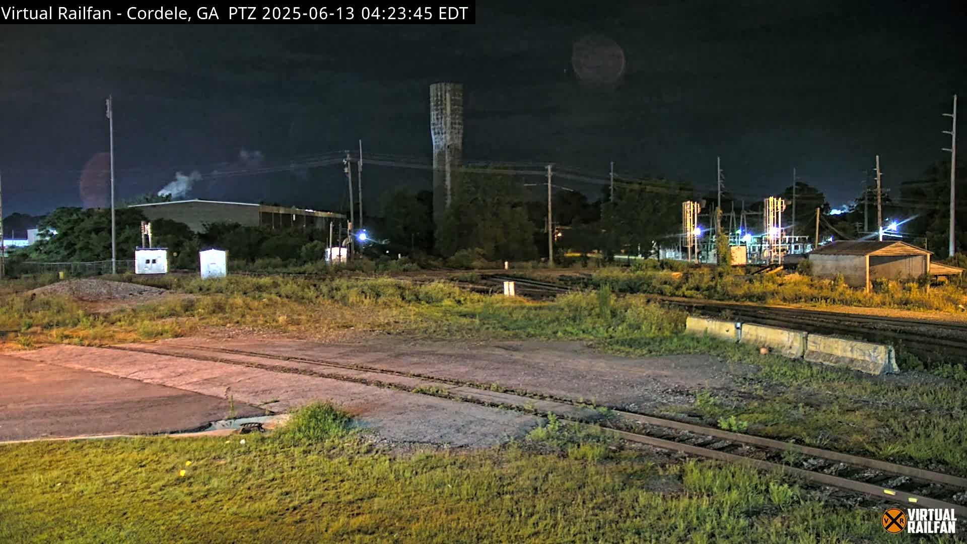 At night, a nighttime view shows a railway switching yard with a tall tower, industrial buildings, and overgrown vegetation under a dark sky.