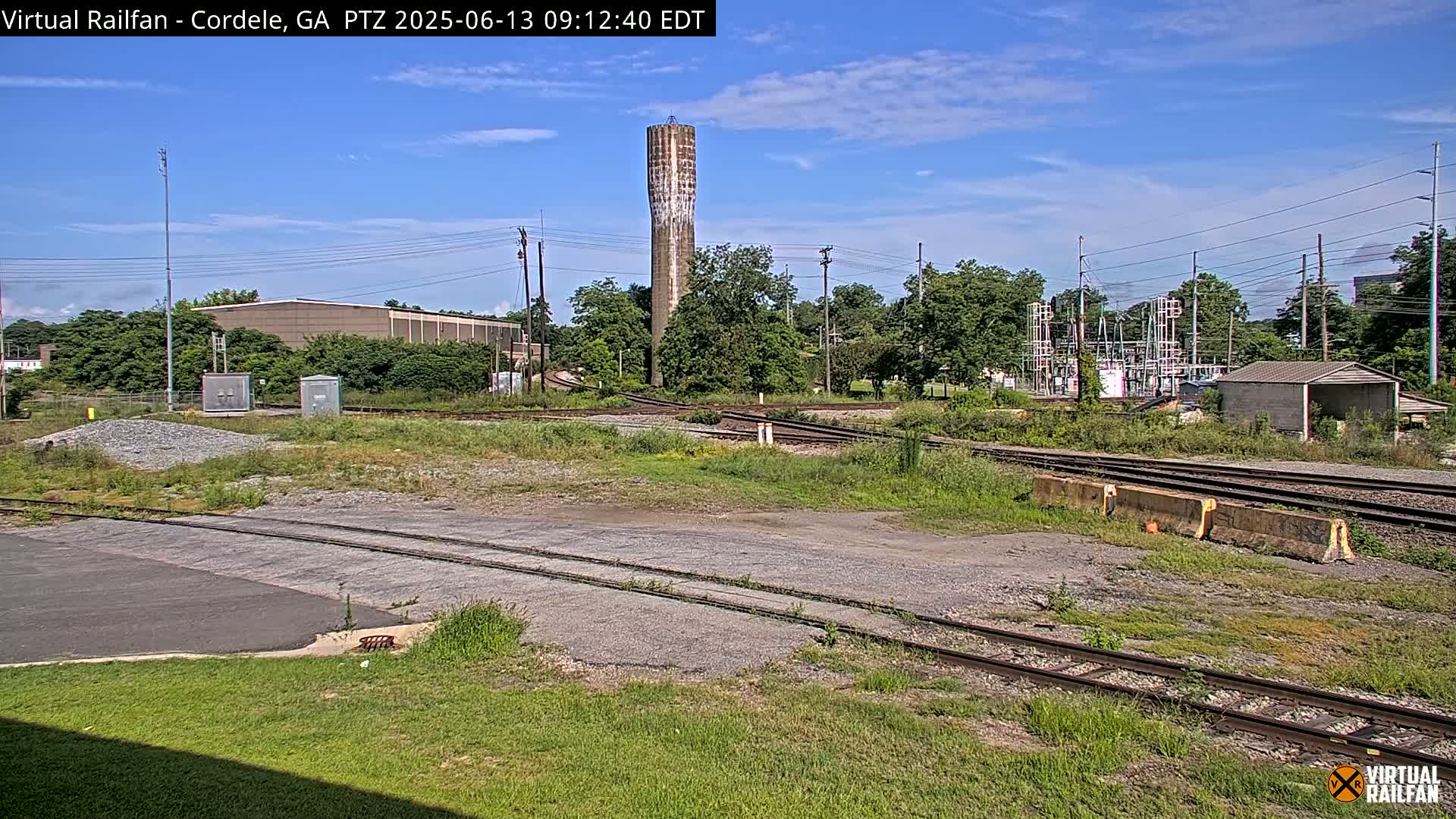 A railroad switching yard with a tall, slender water tower in the background under a mostly sunny sky.