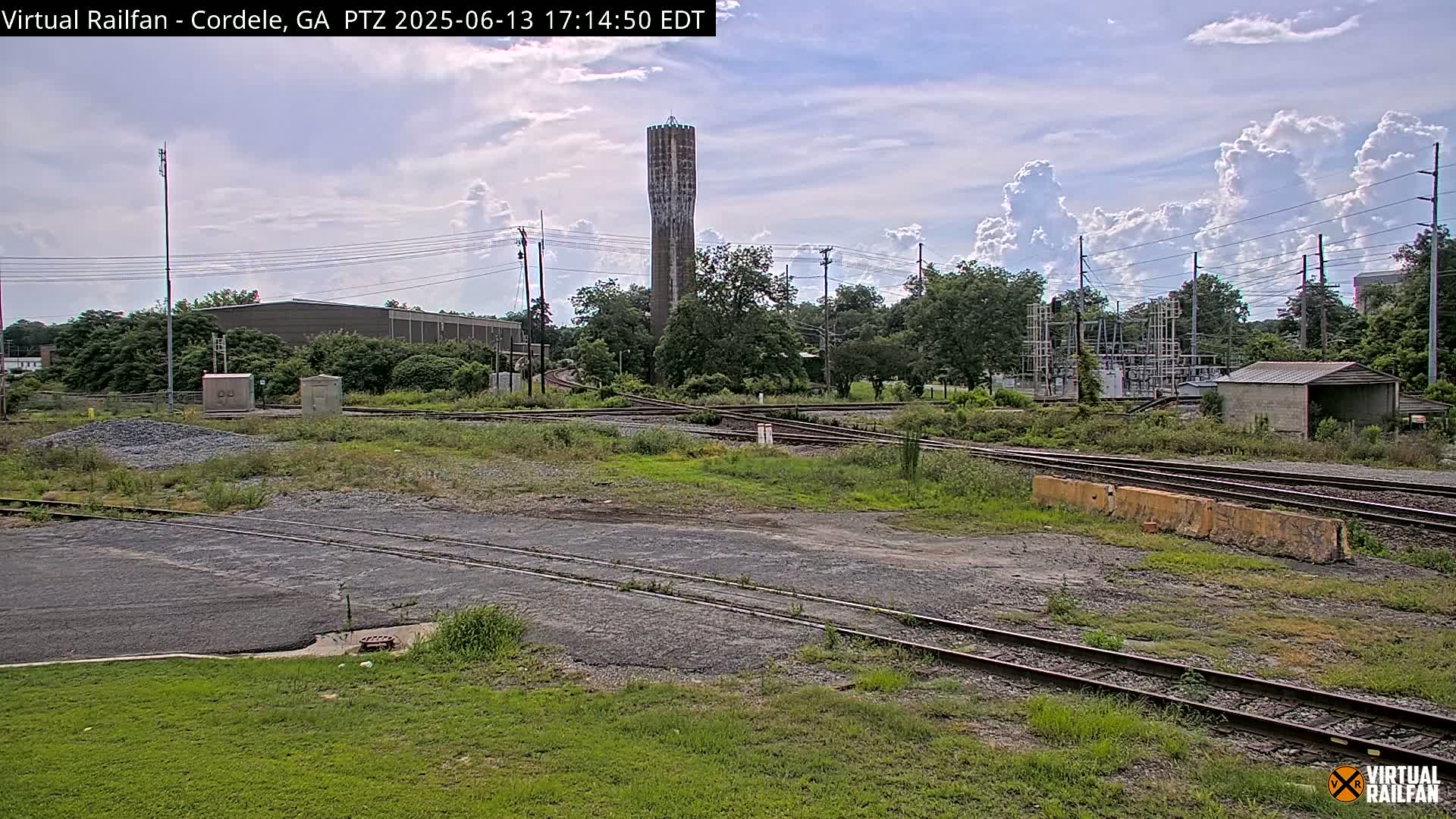 A tall, slender water tower is visible in the distance beyond a railway switching yard under a partly cloudy sky.