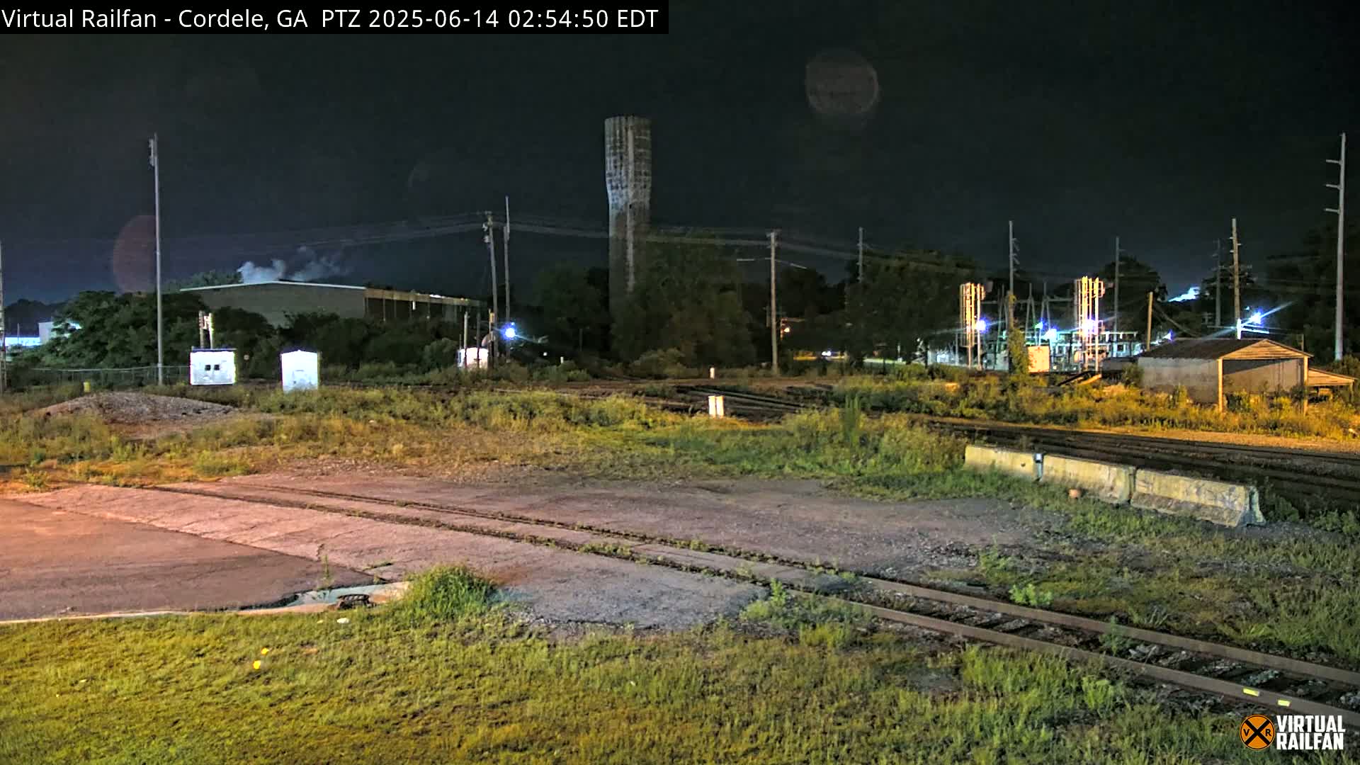 A tall, slender water tower is visible in the distance beyond a railway switching yard under a partly cloudy sky.