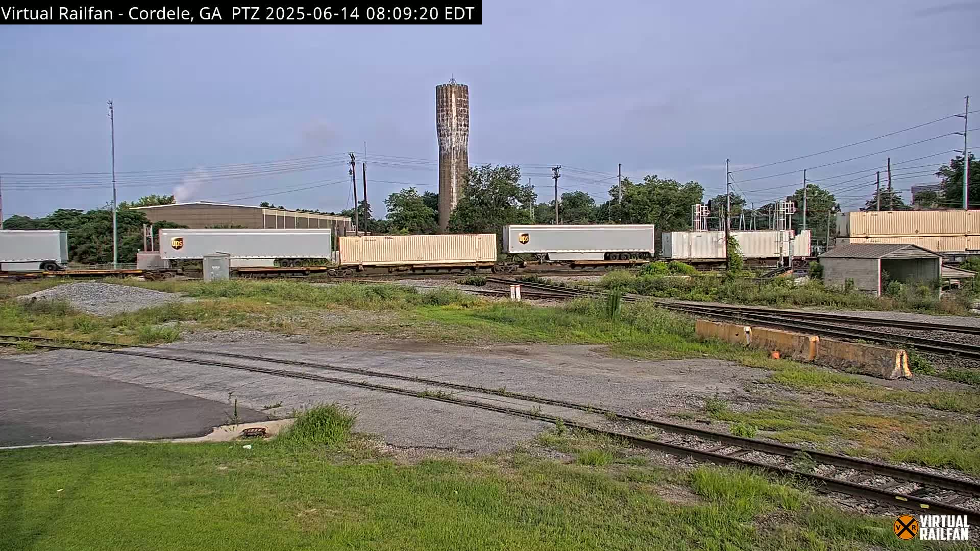 Railroad tracks and several UPS shipping containers sit on a rail yard next to a tall water tower under a mostly clear sky.