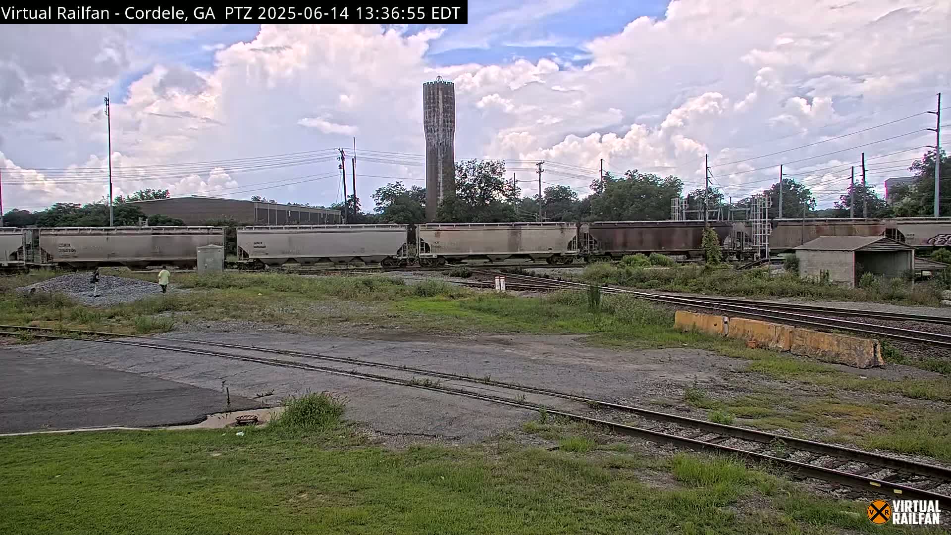A partly cloudy day shows a rail yard with several freight cars, a water tower in the background, and a person walking near a pile of gravel.